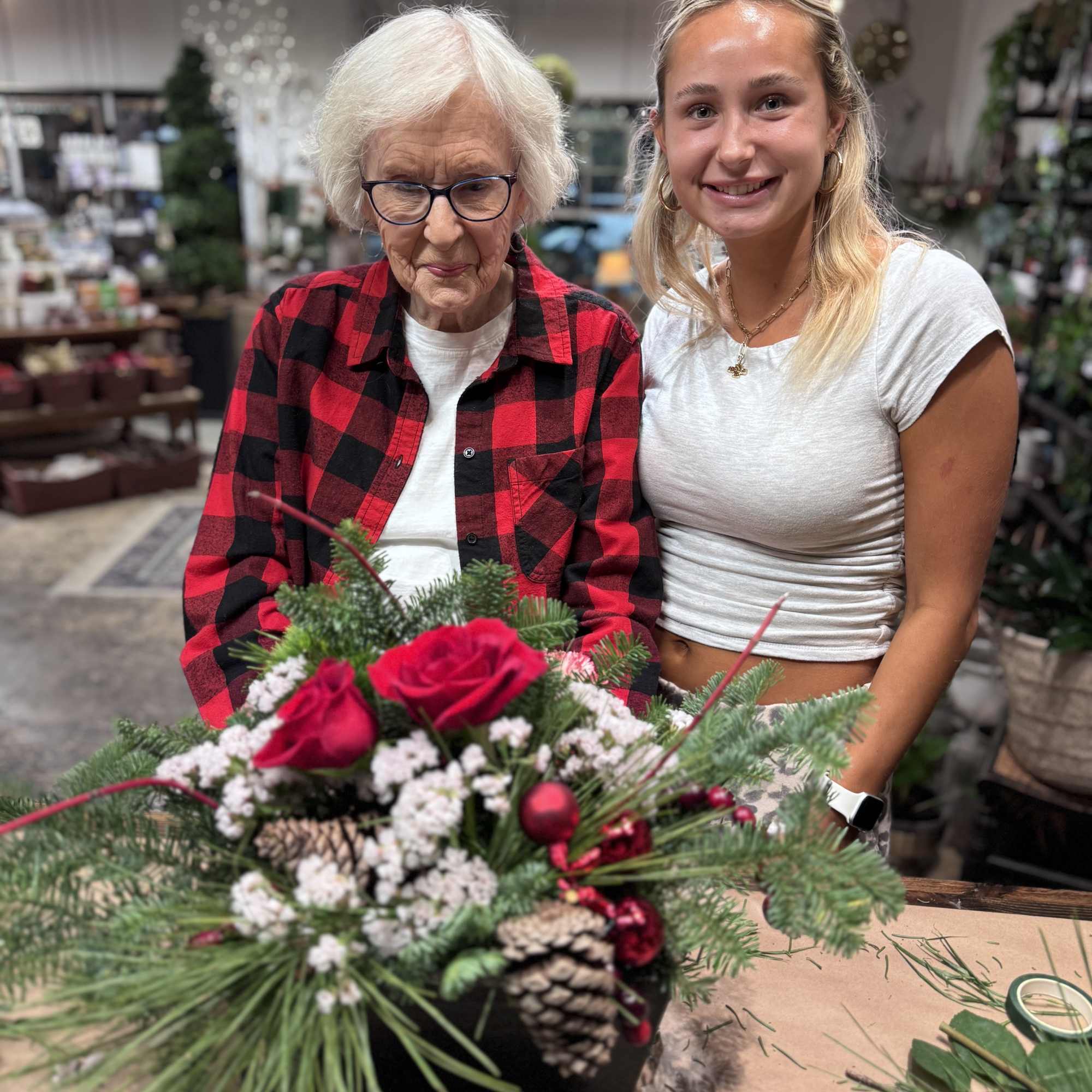 Two women hold a red rose and pine arrangement in a black container