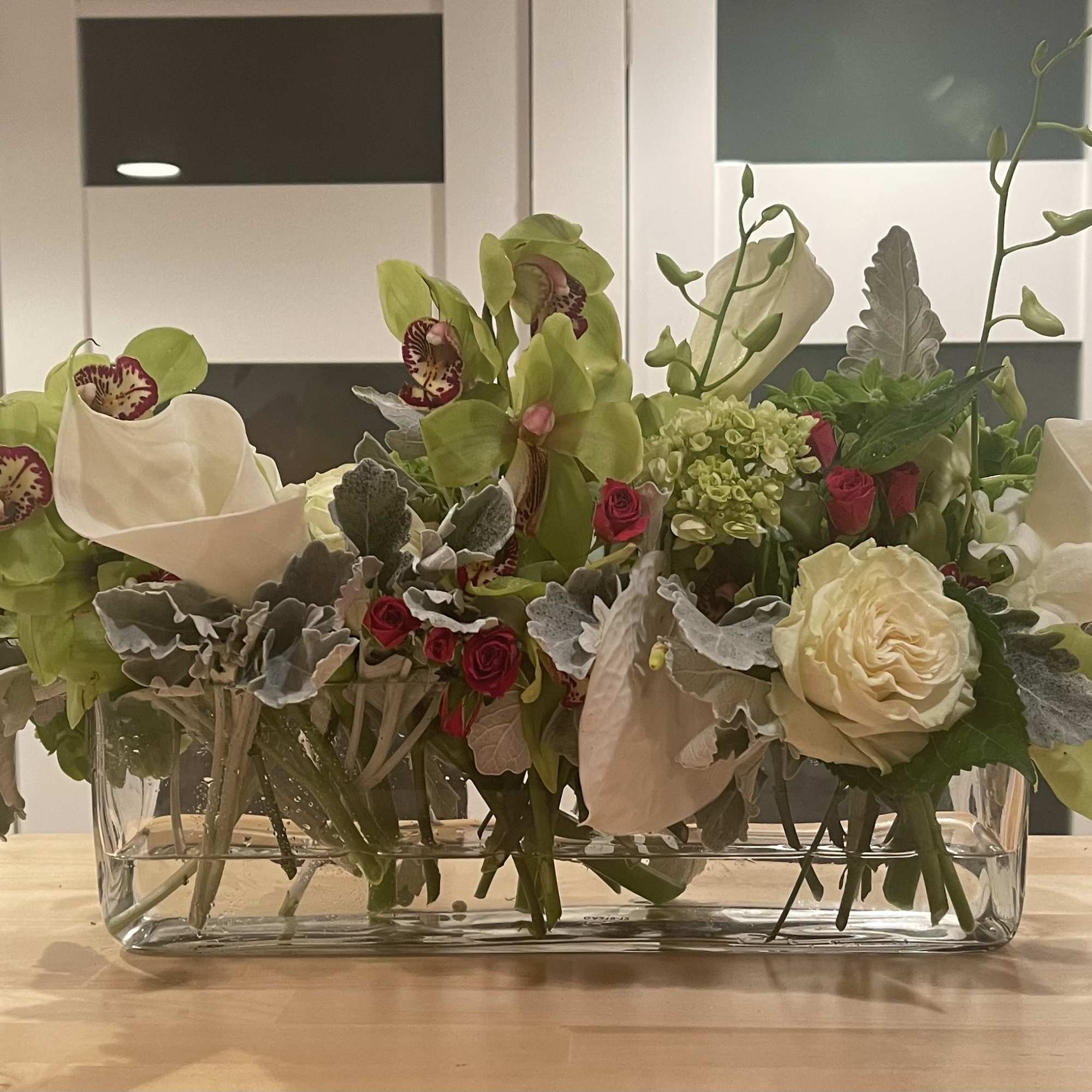 Low floral arrangement in a clear rectangular glass vase with orchids, roses, and calla lilies