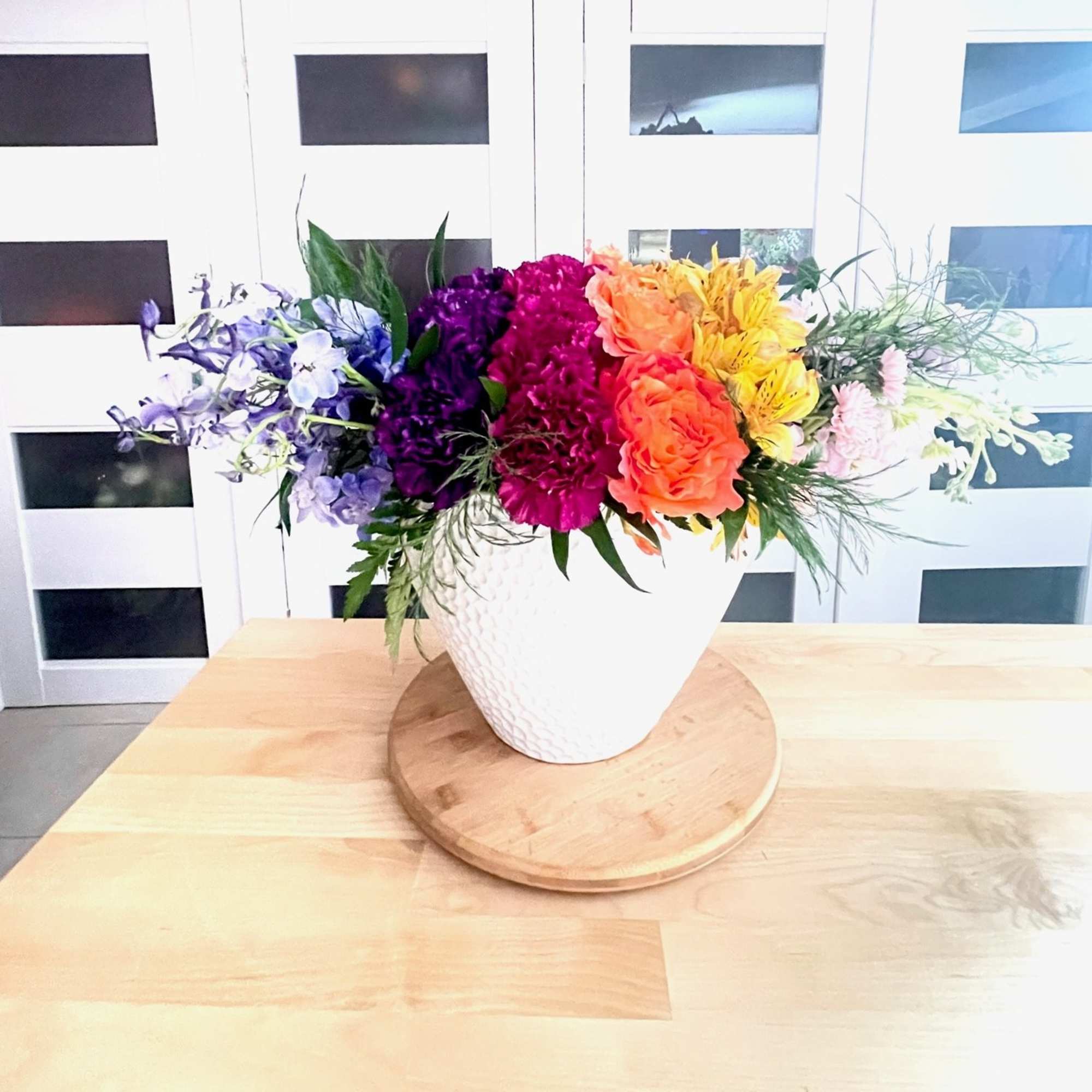 Rainbow floral arrangement in a white vase on a wooden tray