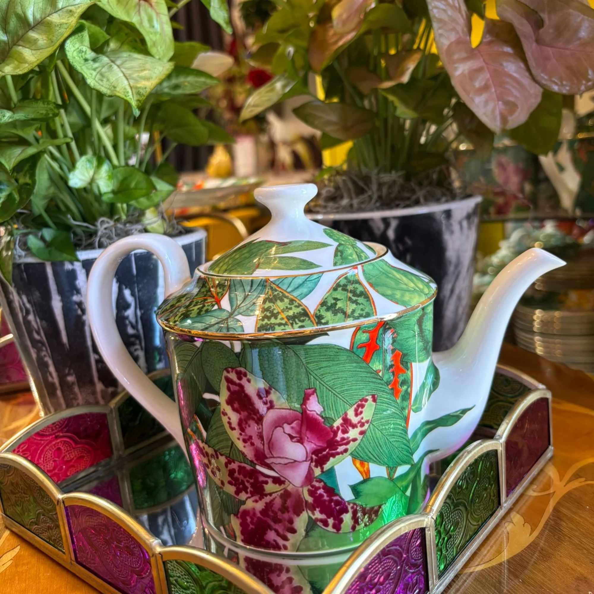 Decorative teapot with floral pattern on a mirrored tray, with potted plants behind it