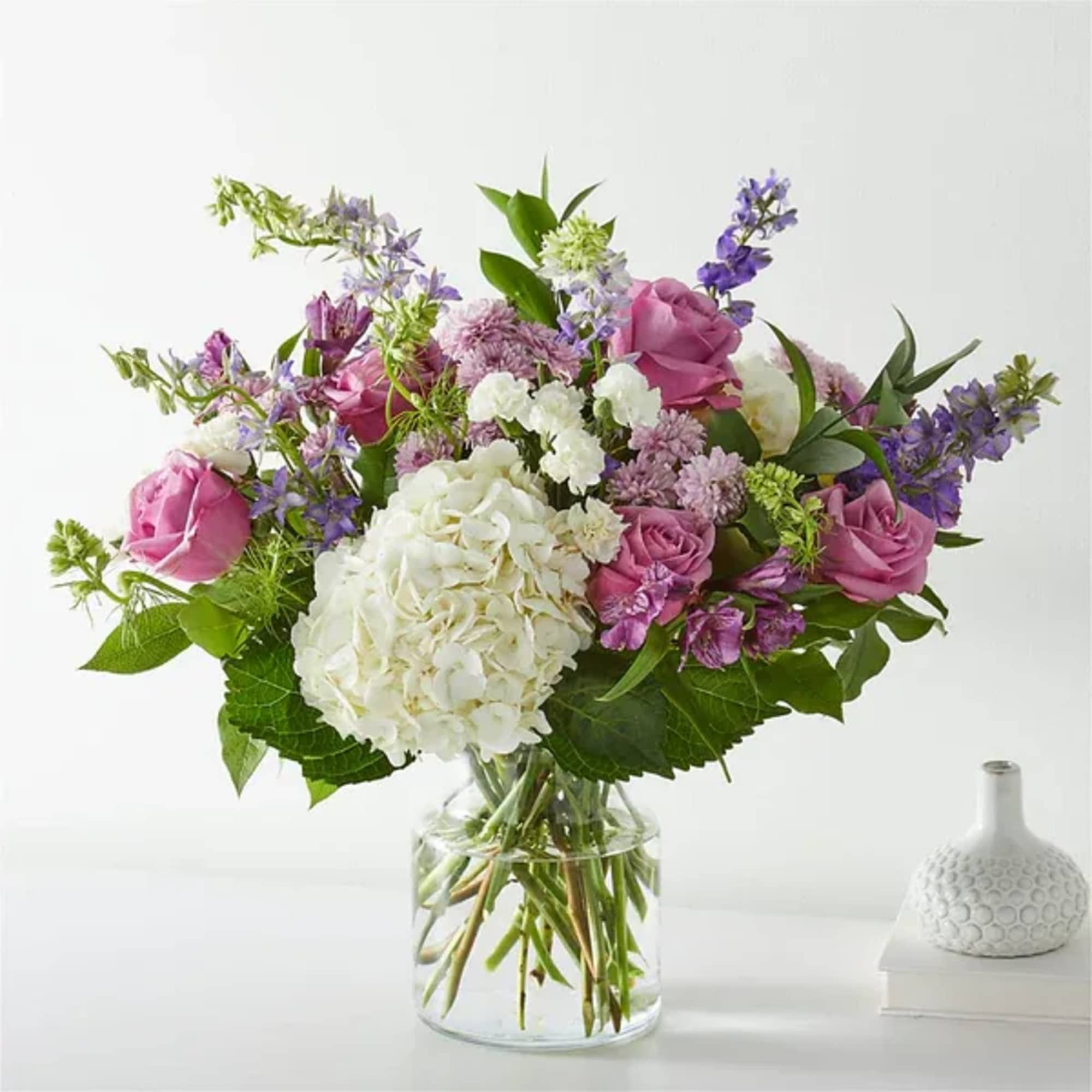 Mixed arrangement of pink roses, white hydrangea, and purple flowers in a clear glass vase