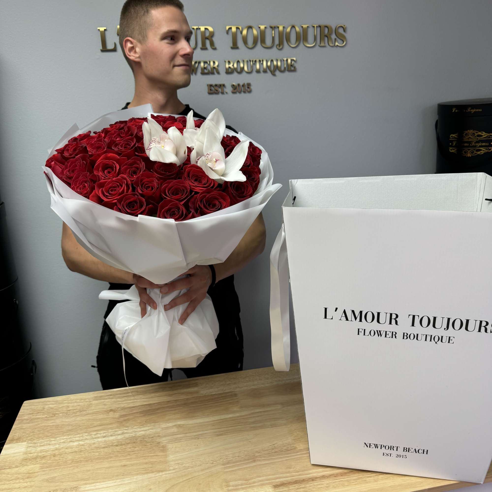 Large bouquet of red roses with white orchids wrapped in white paper, held by a person indoors.