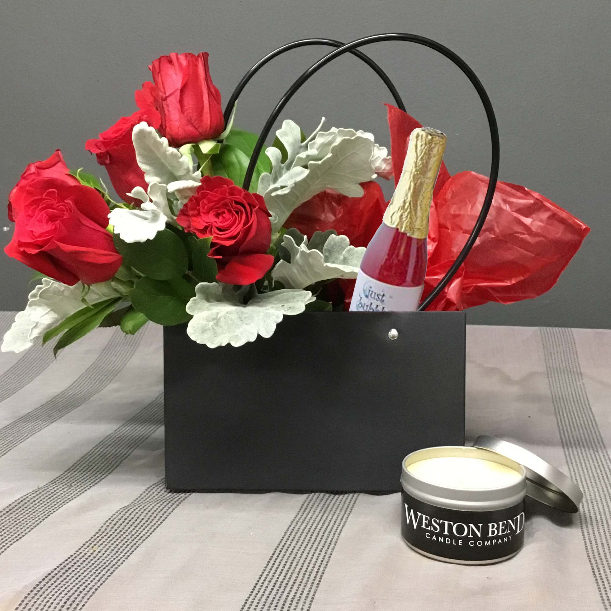 Red roses in a black gift bag with a candle and a small bottle of sparkling drink