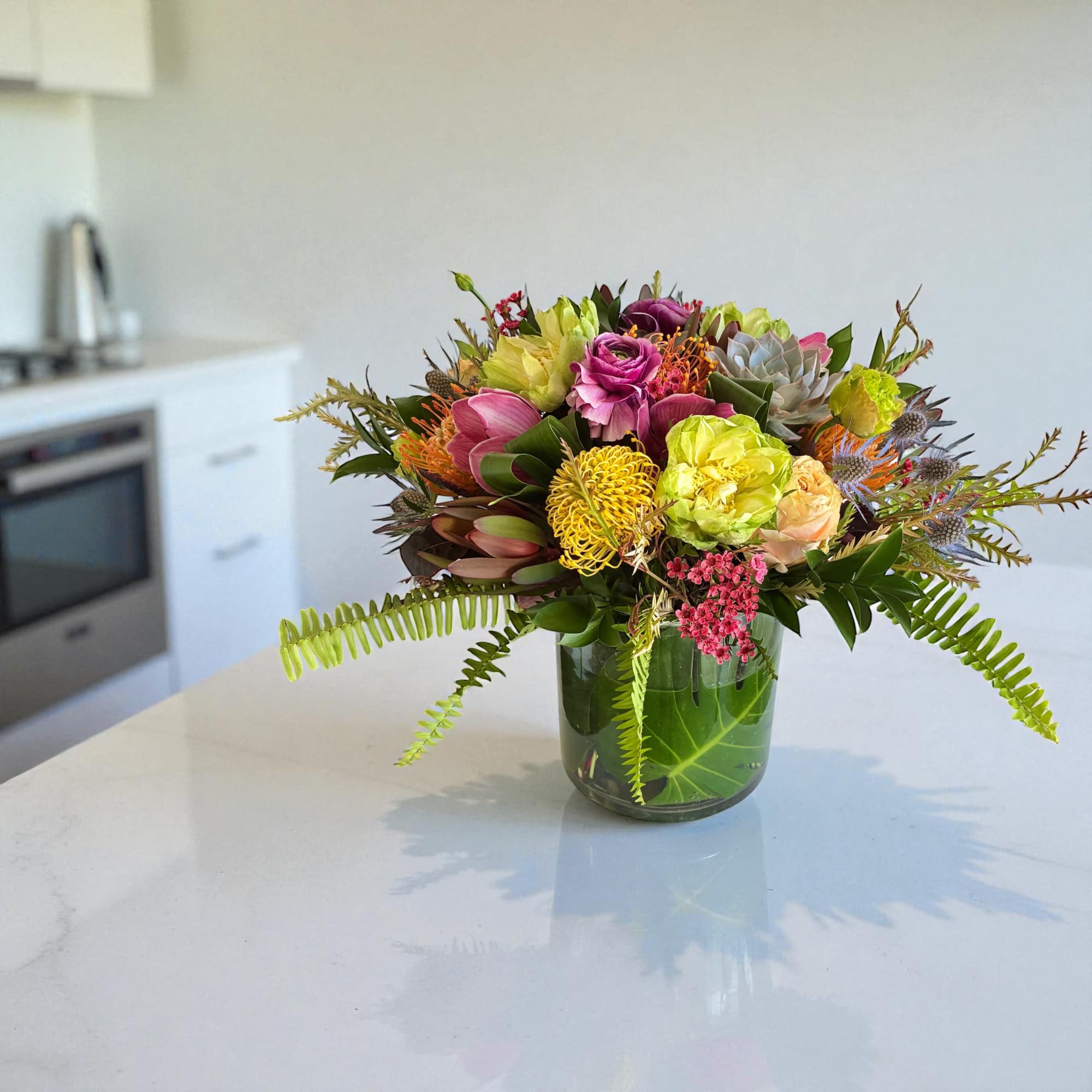 Bright mixed bouquet with protea, roses, and tropical greenery in a clear glass vase on a countertop