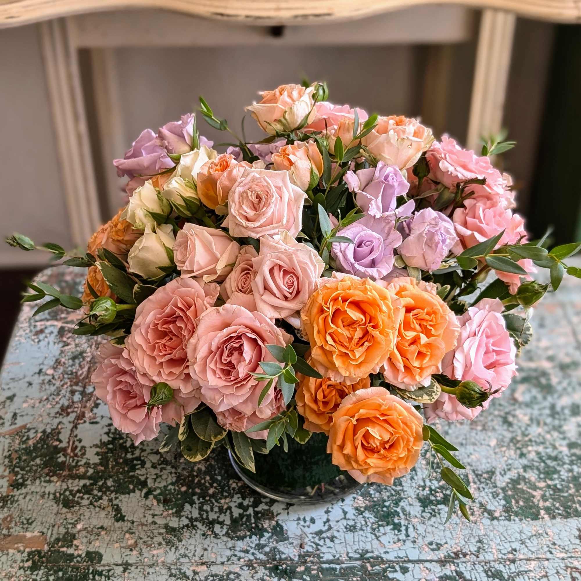 Low vase of pink, peach, and lavender roses arranged in a rounded bouquet on a rustic painted table