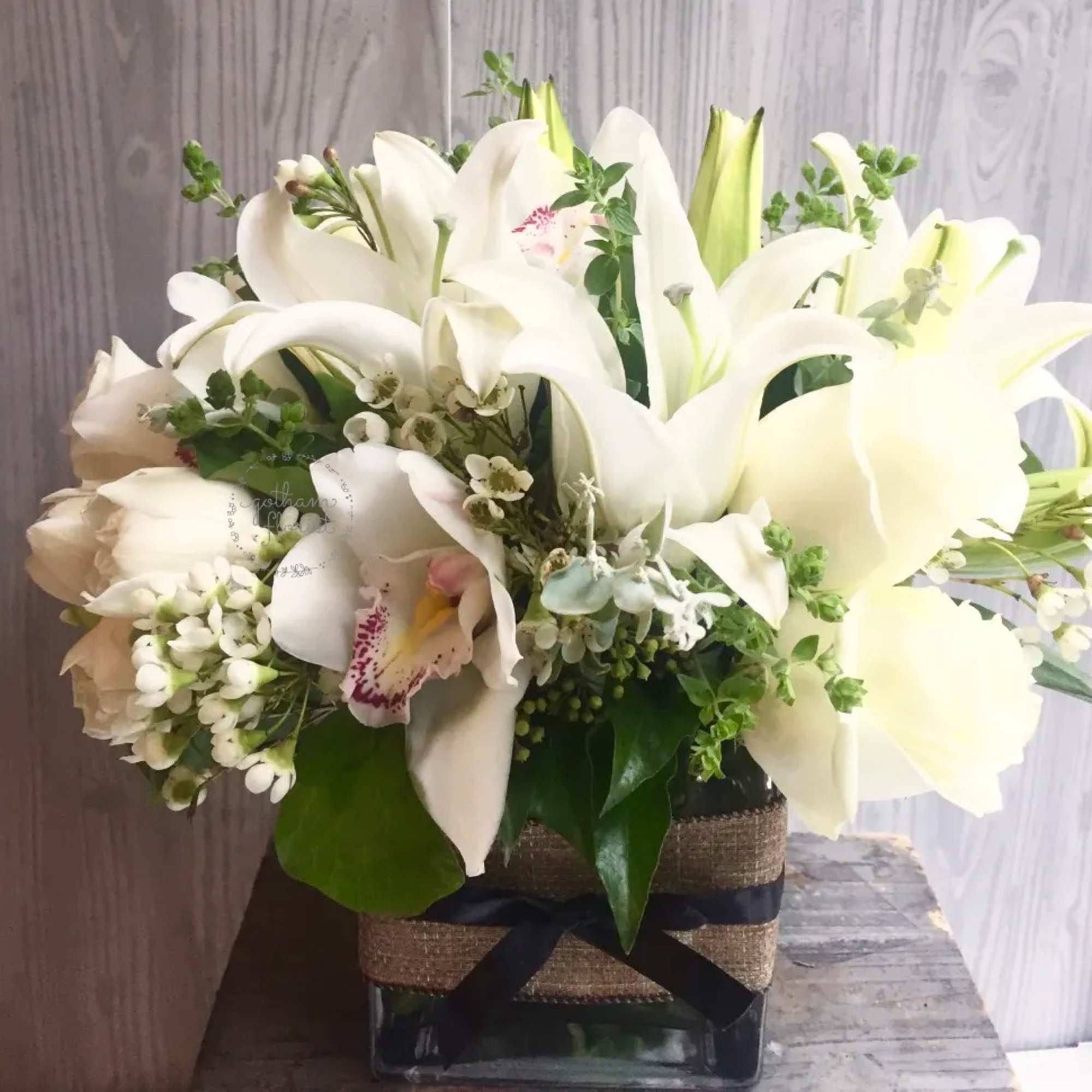 White lilies and orchids arranged in a square glass vase with a ribbon wrap.