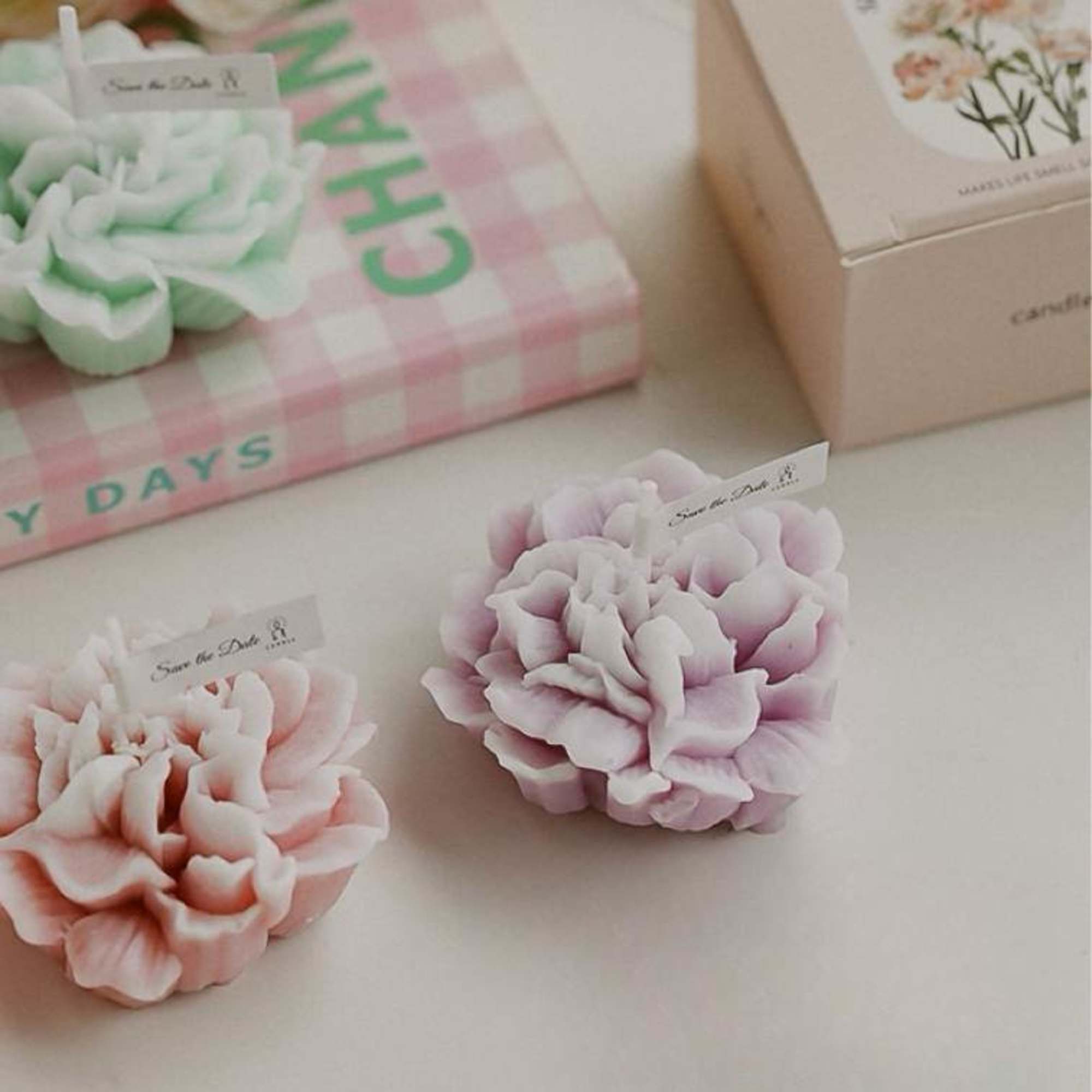 Three pastel flower-shaped candles on a tabletop, with gift boxes in the background