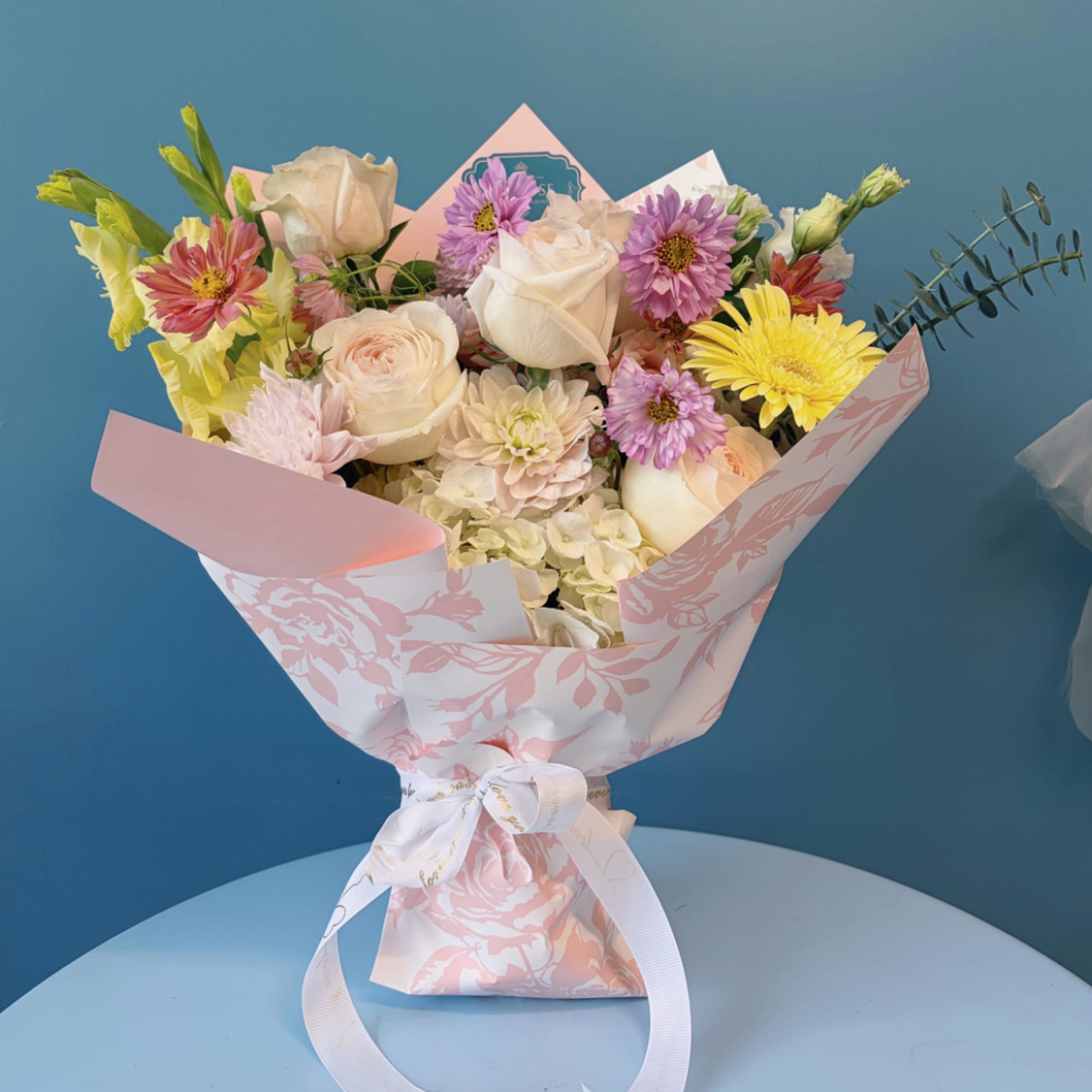 Bouquet of mixed pastel flowers wrapped in pink paper with a white ribbon