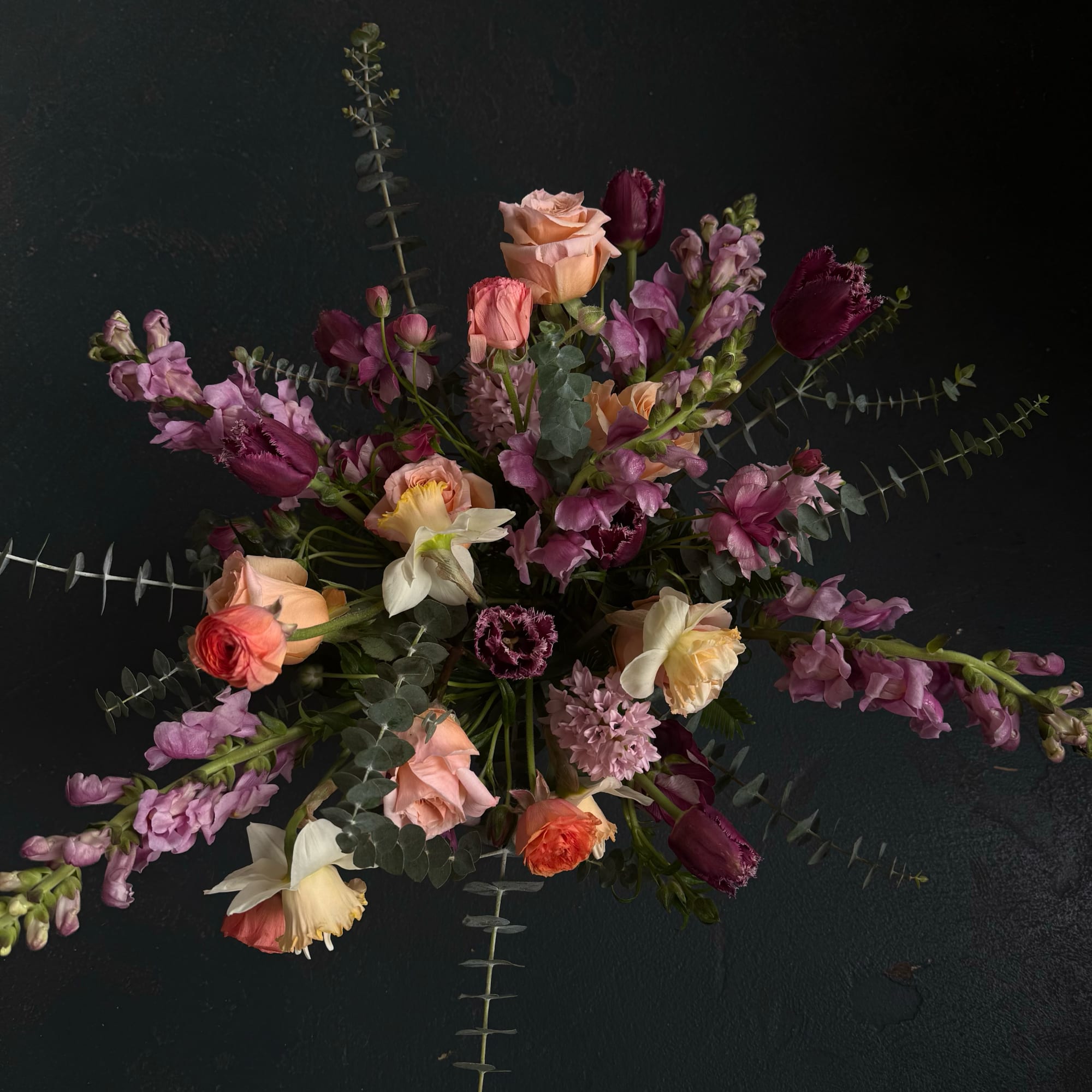 Moody arrangement of peach roses, white daffodils, and purple tulips on a dark background