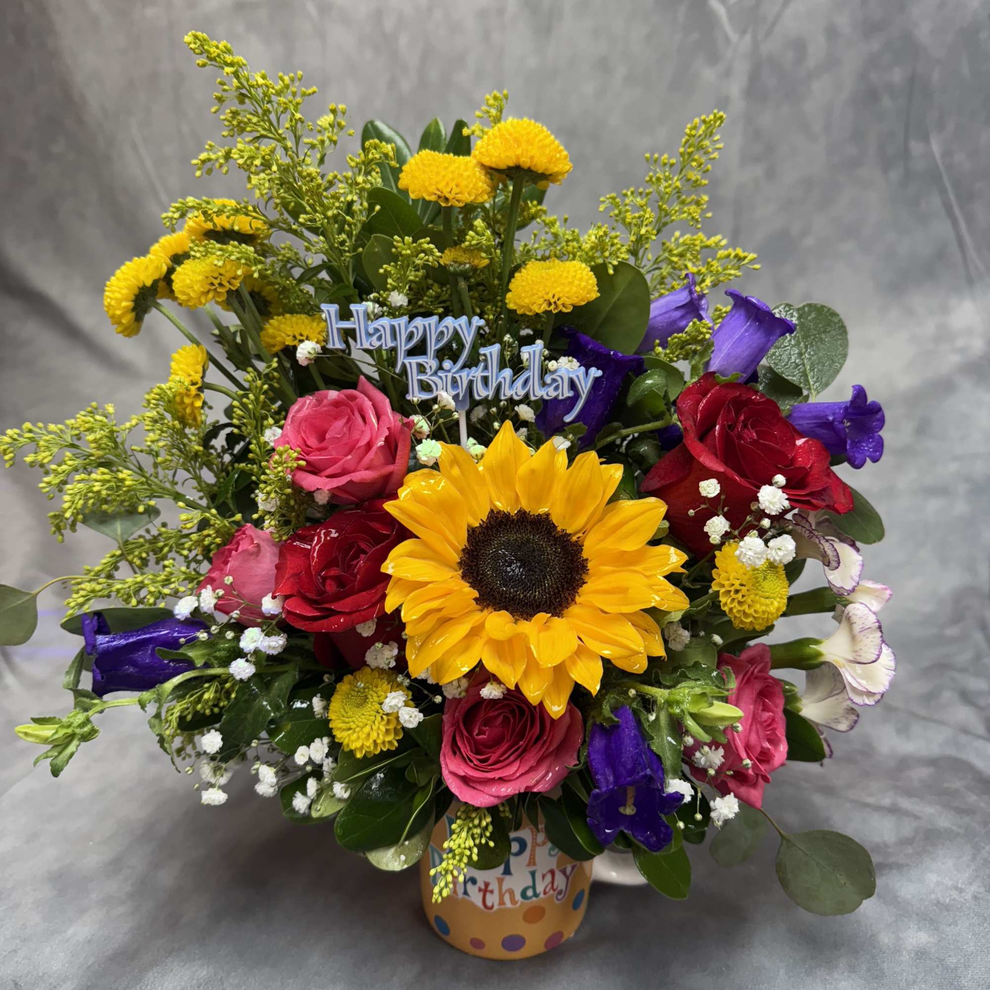 Mixed bouquet with a sunflower, roses, and purple blooms in a birthday container