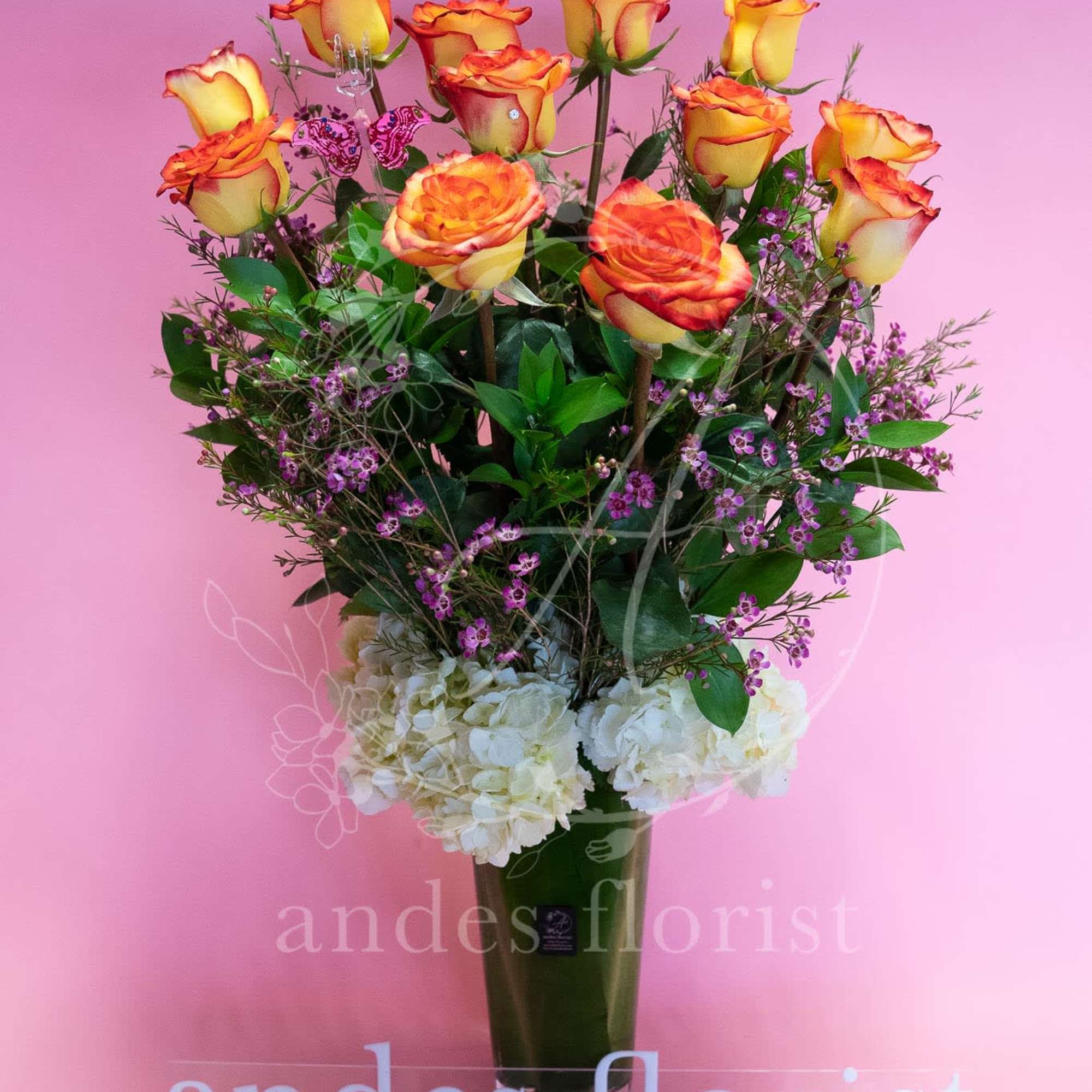 Orange-yellow roses in a tall vase with white hydrangeas and purple filler flowers