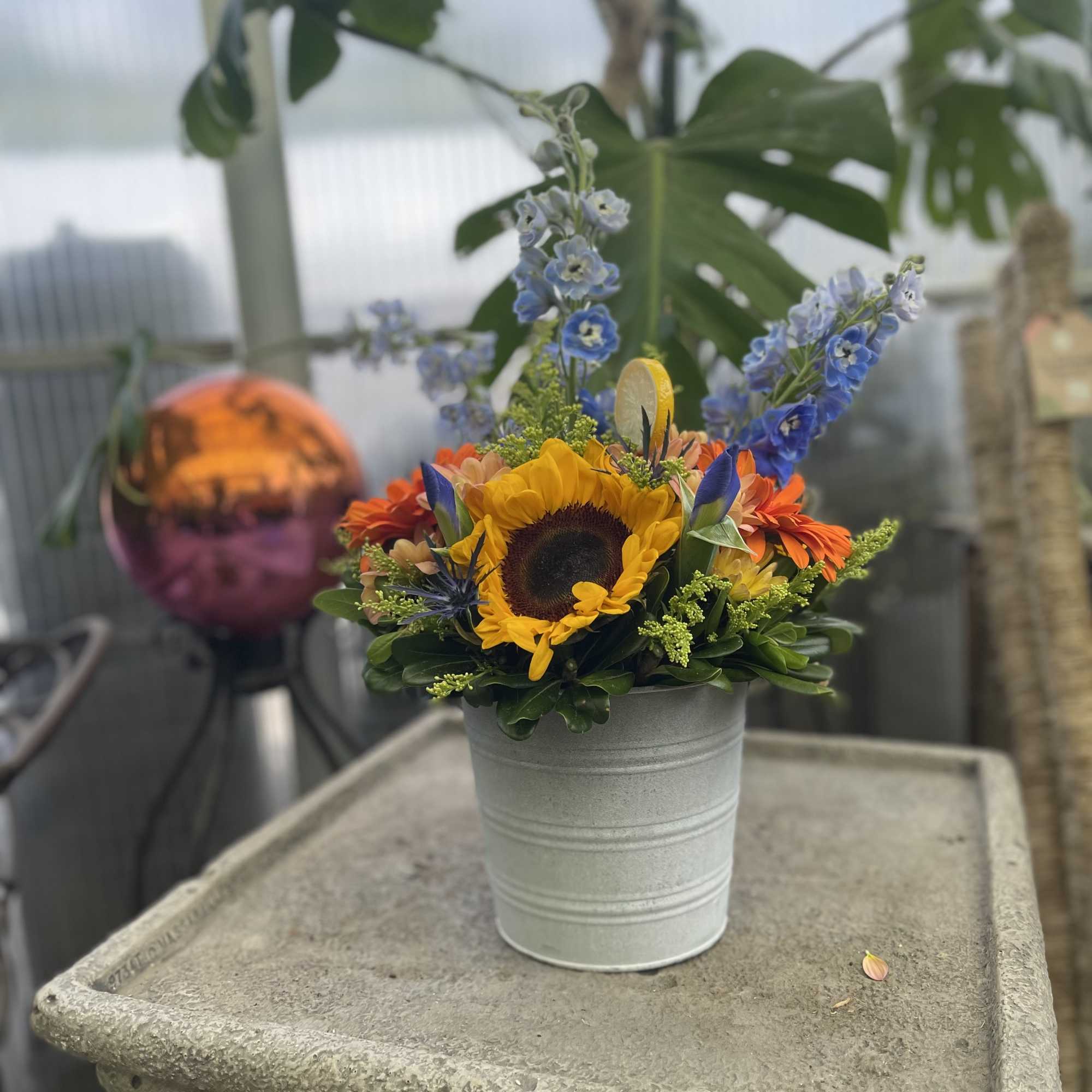 Sunflower arrangement in a small white metal pot with blue and orange blooms