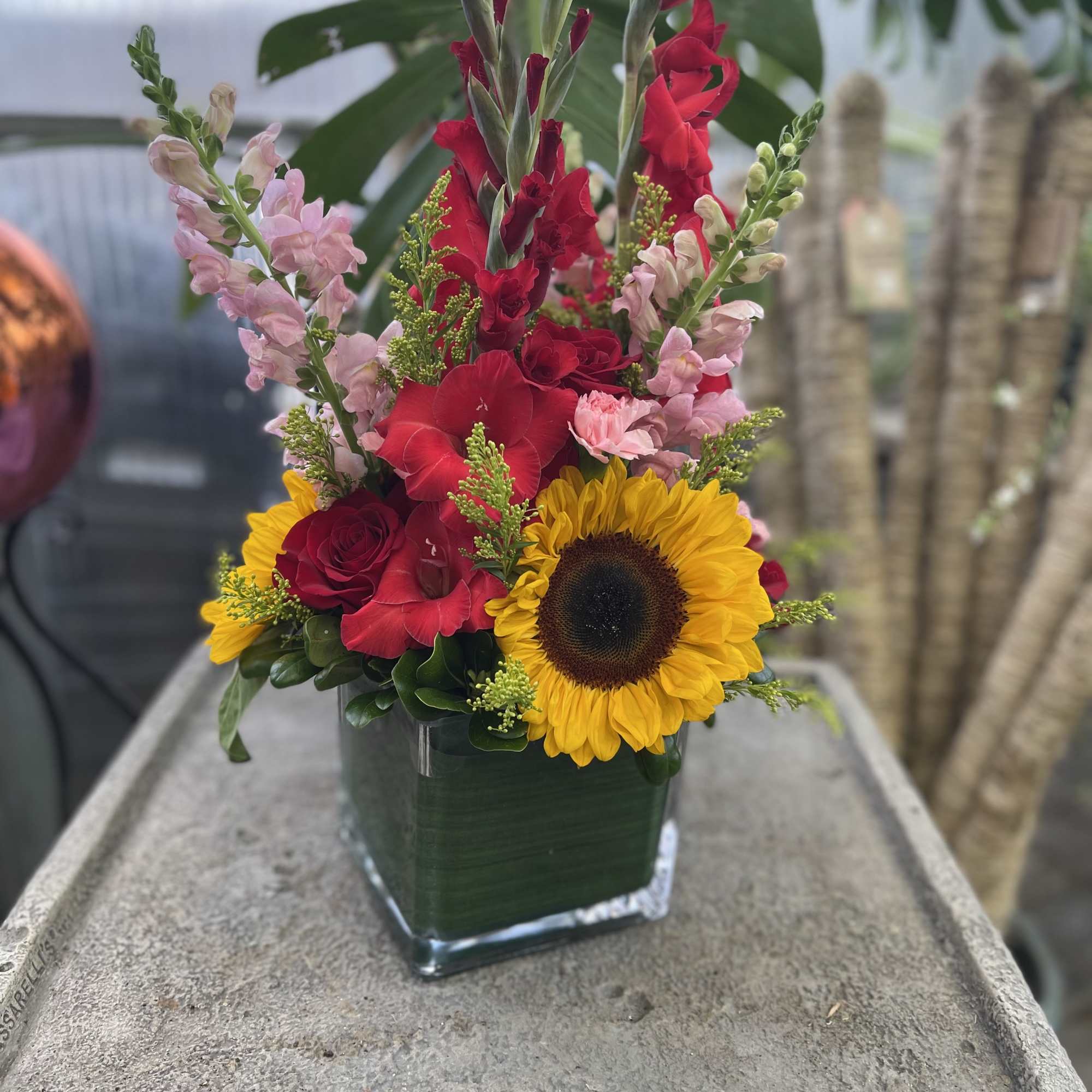Tall floral arrangement with sunflowers, red gladiolus, and pink snapdragons in a square vase