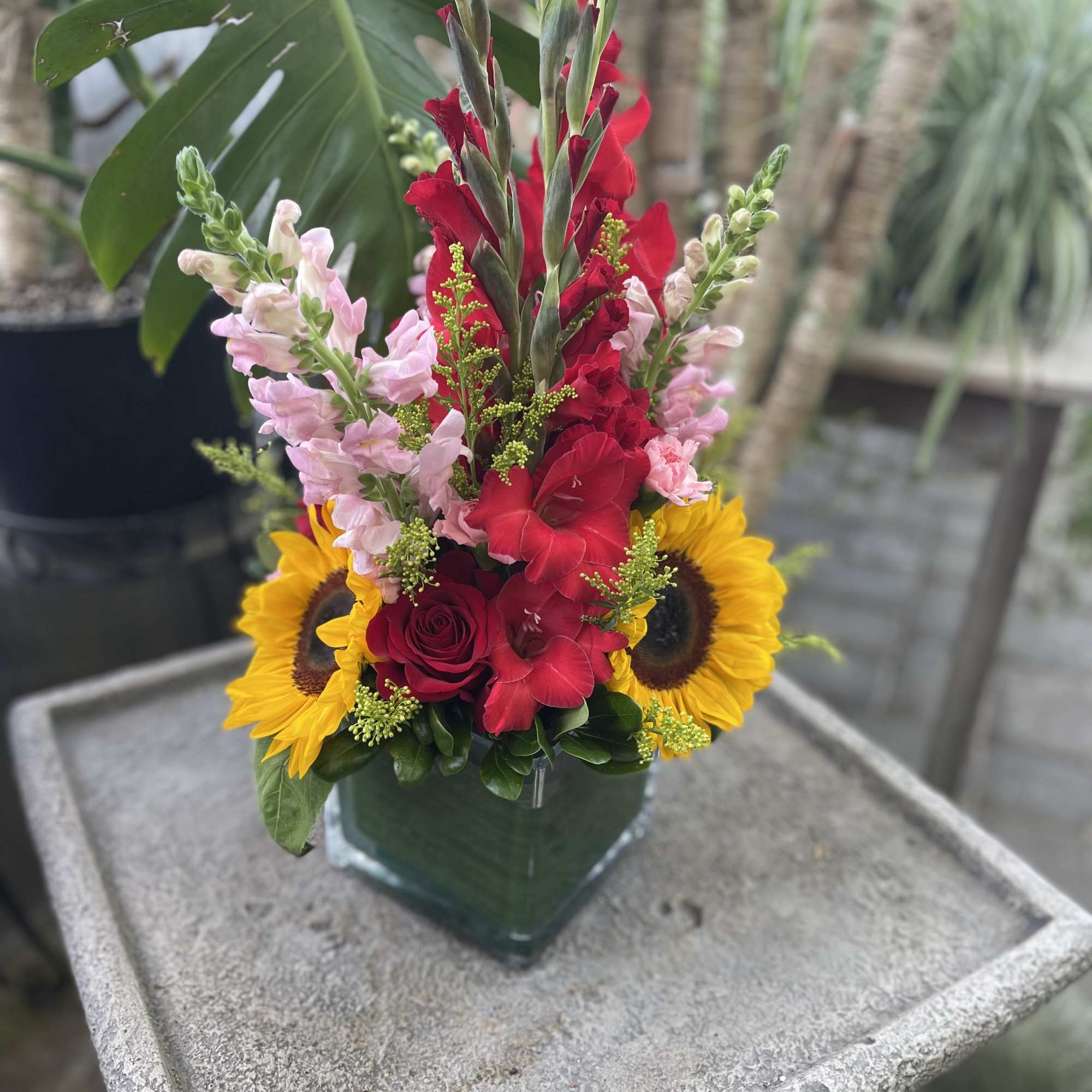 Bright mixed bouquet in a square glass vase with sunflowers, red gladiolus, and pink snapdragons