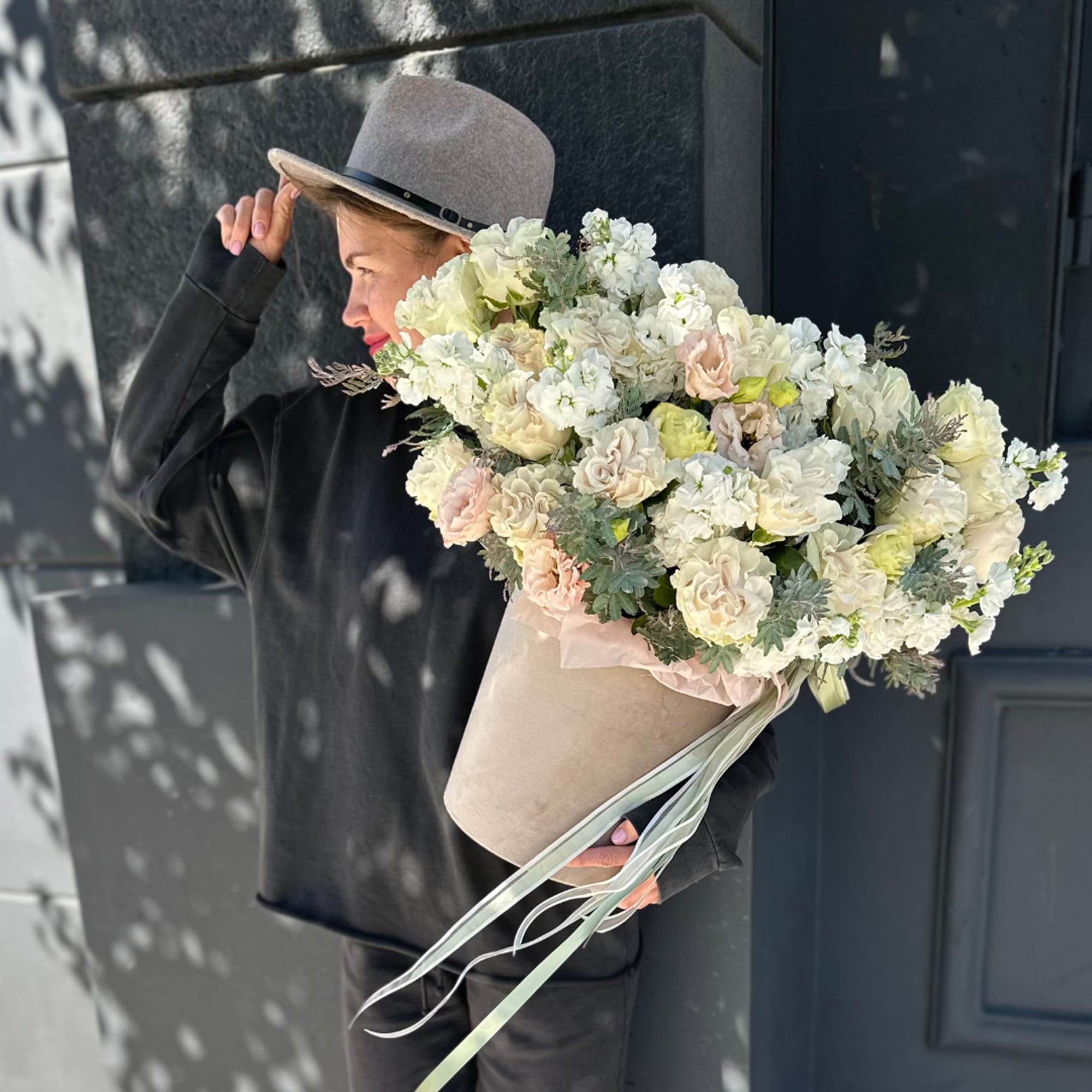 Large bouquet of white and blush flowers in a beige wrap, held by a person in a hat.