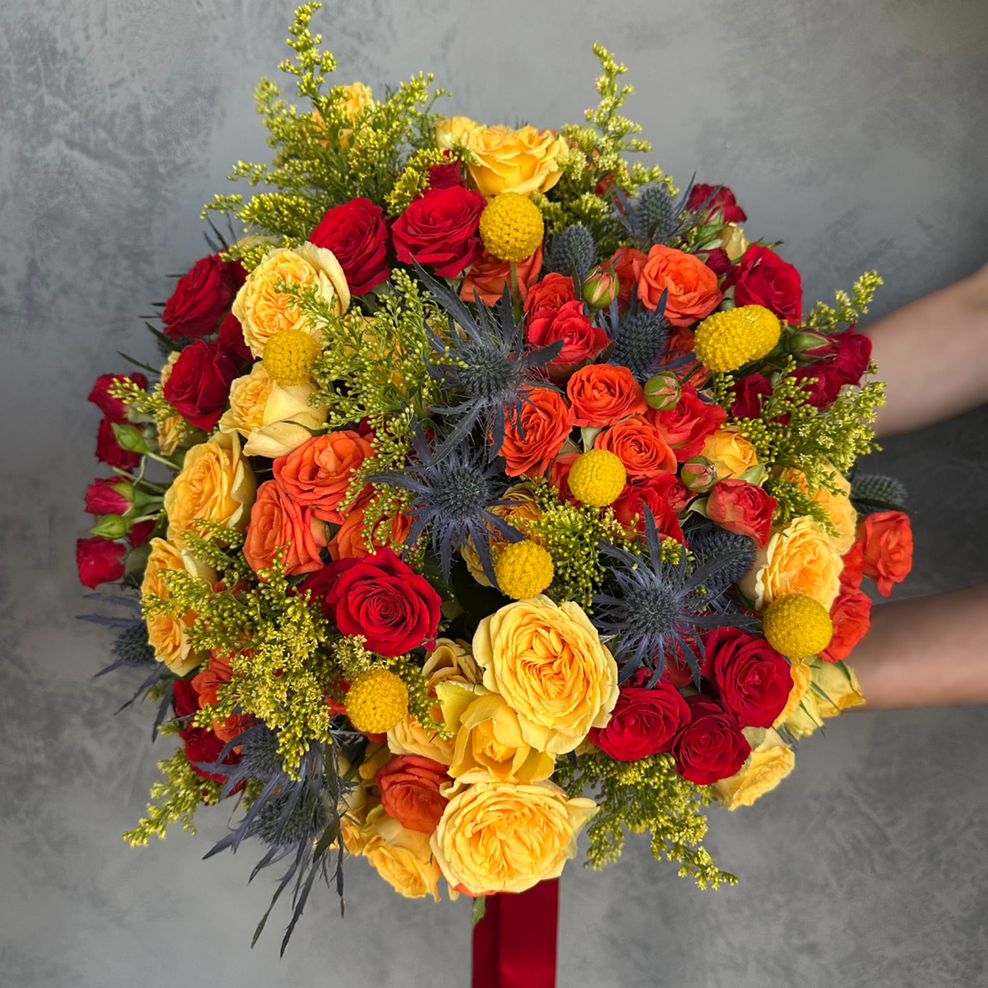 Round bouquet of red, orange, and yellow roses with blue thistle accents