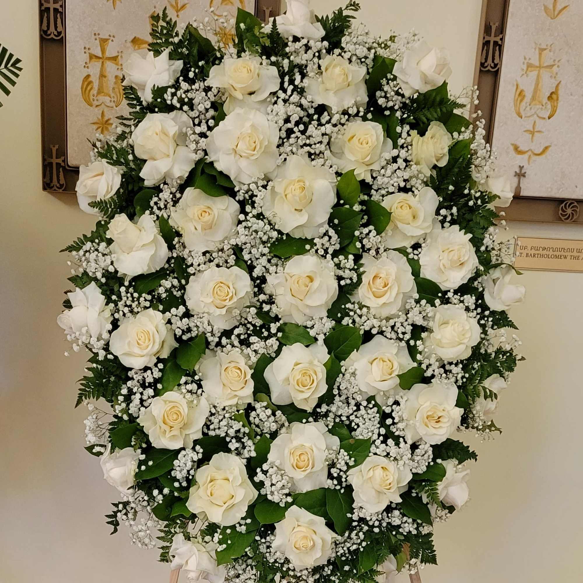 Oval standing spray of white roses and baby's breath on an easel with a black dedication sign.
