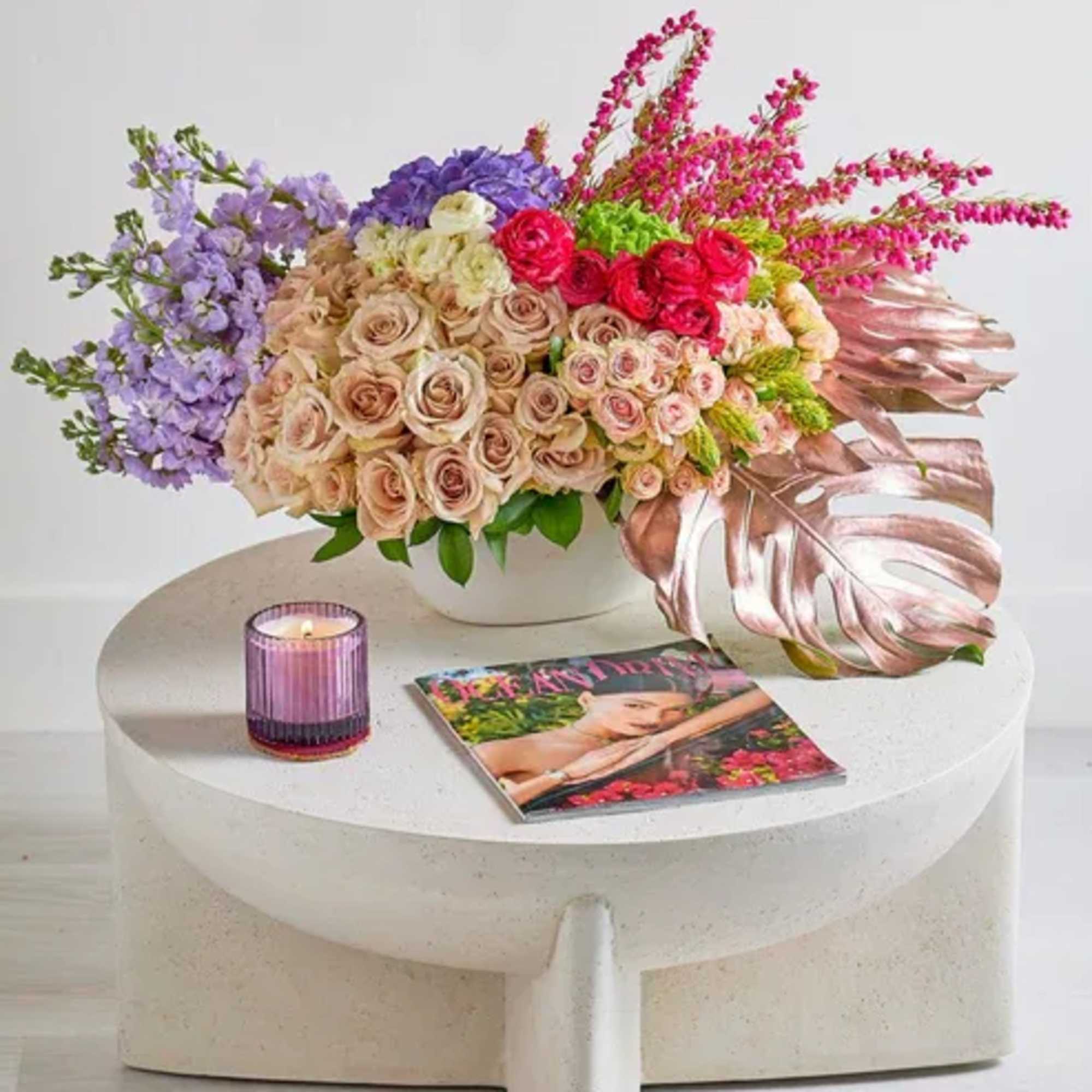 Mixed bouquet of roses and purple flowers in a white vase with a candle and magazine
