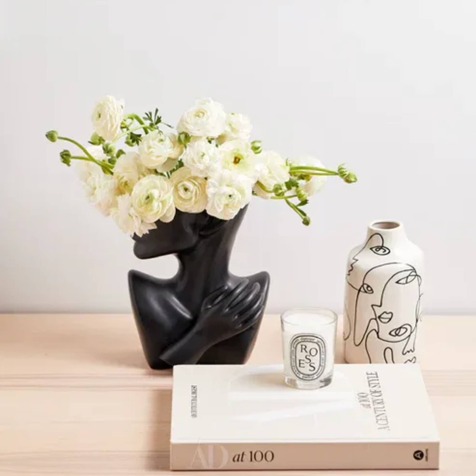 White ranunculus arrangement in a black sculptural vase with a candle and decorative vase