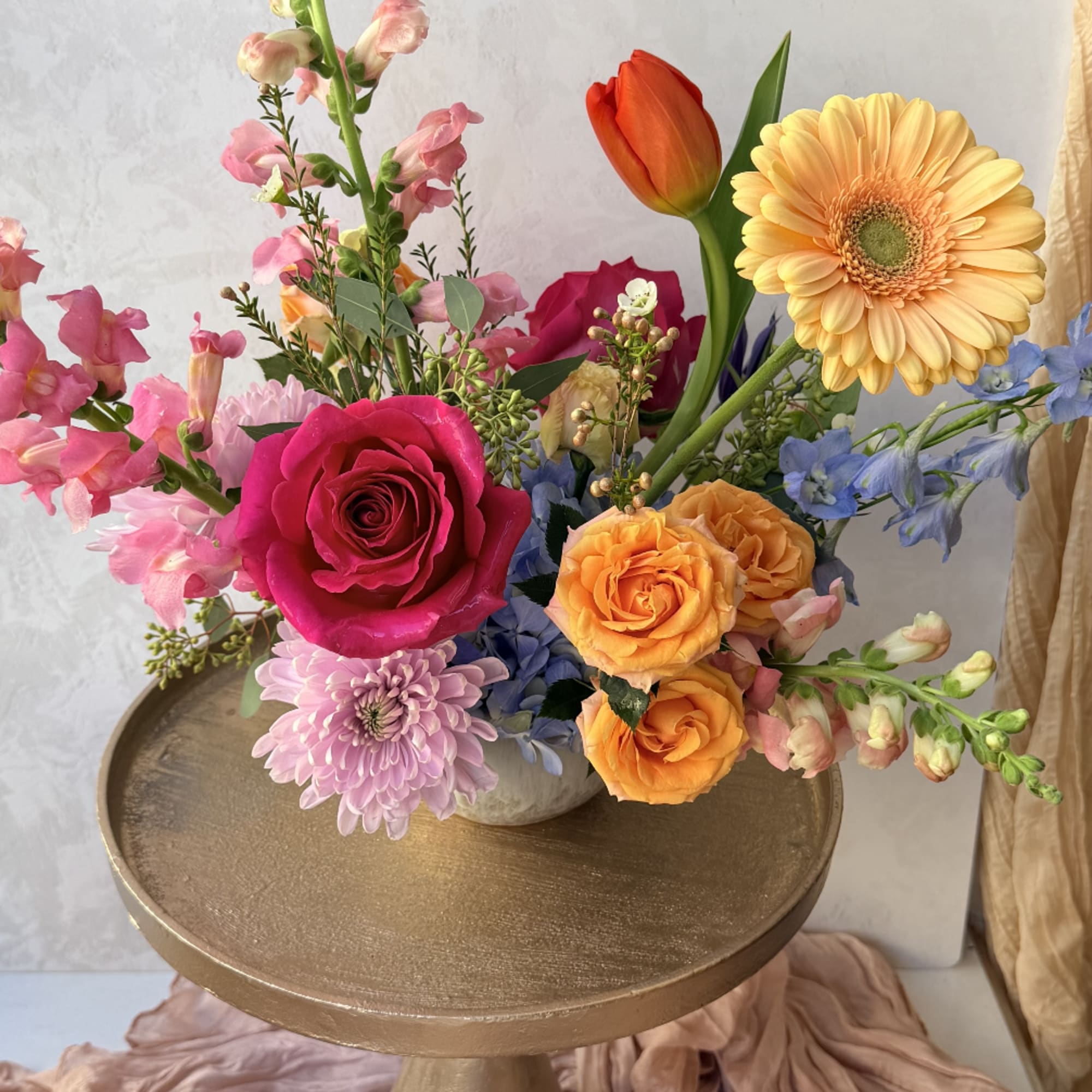 Colorful mixed arrangement of pink, orange, and peach flowers in a low vase on a gold pedestal stand