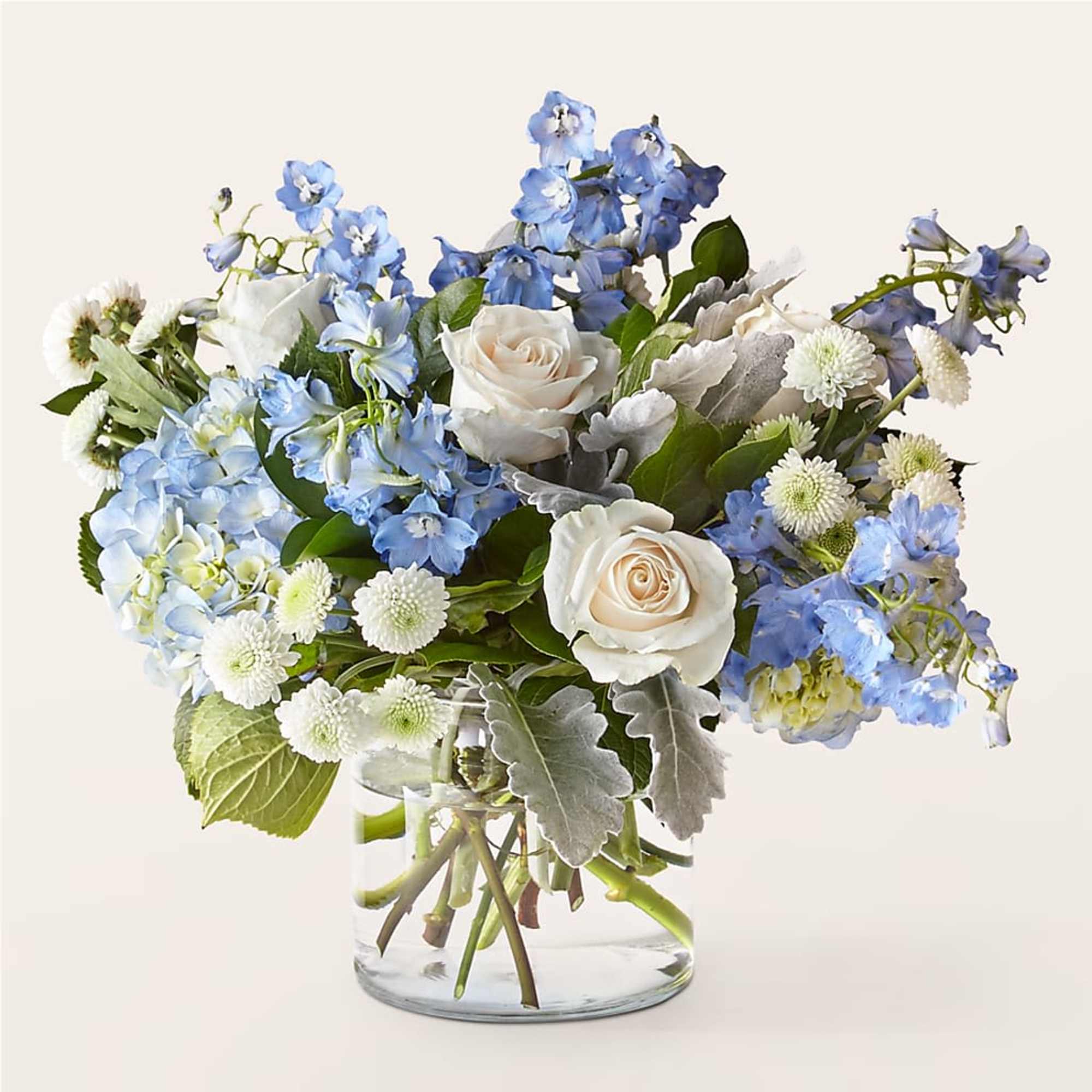 Low glass vase arrangement of white roses, blue delphinium, blue hydrangea, and white button mums