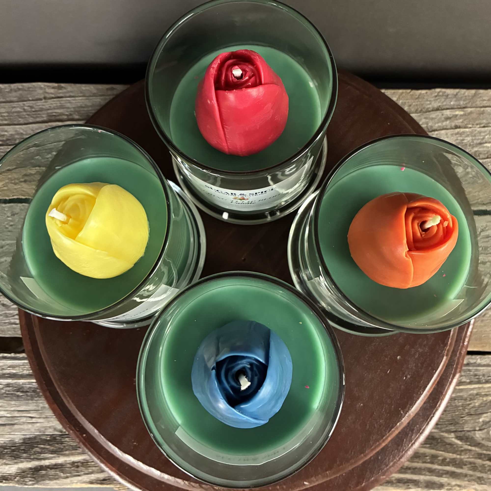 Set of four glass candles with green wax and rose-shaped centers in red, yellow, blue, and orange.