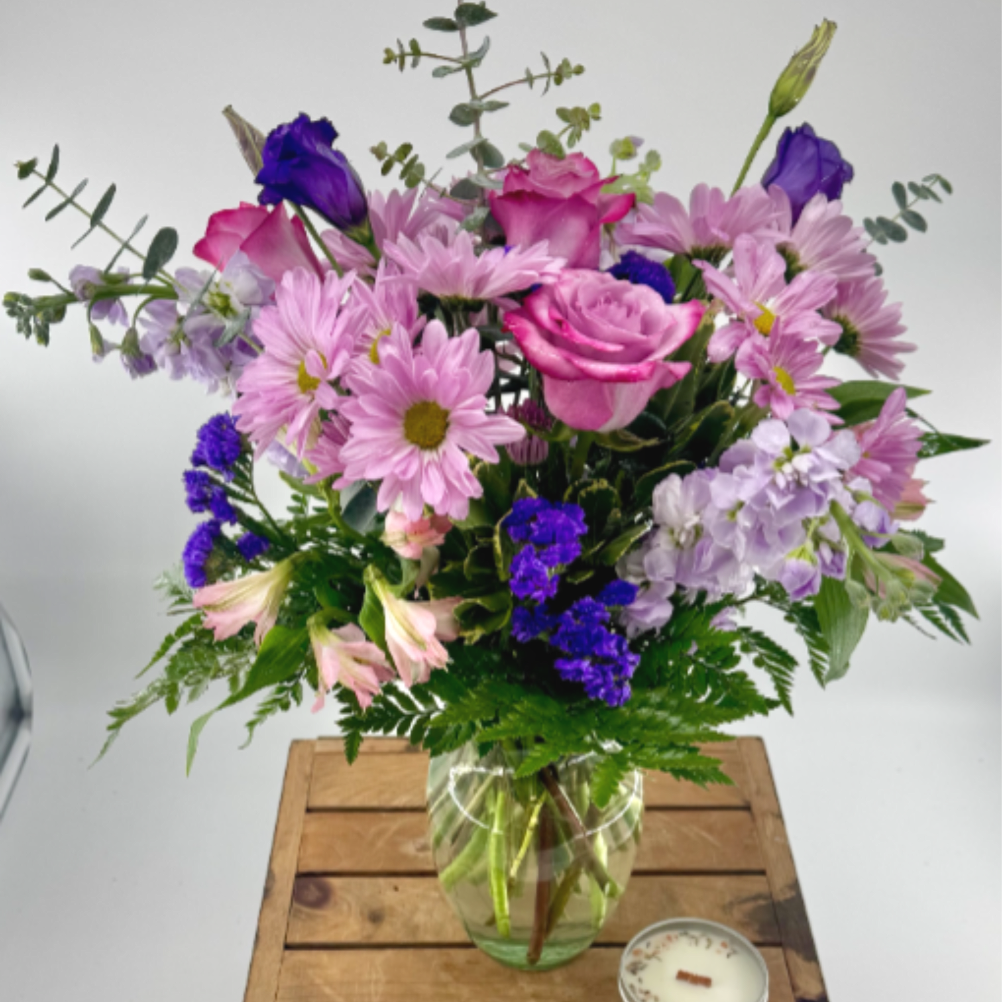 Pink and purple mixed flower bouquet in a clear glass vase on a wood crate with a small tin candle beside it