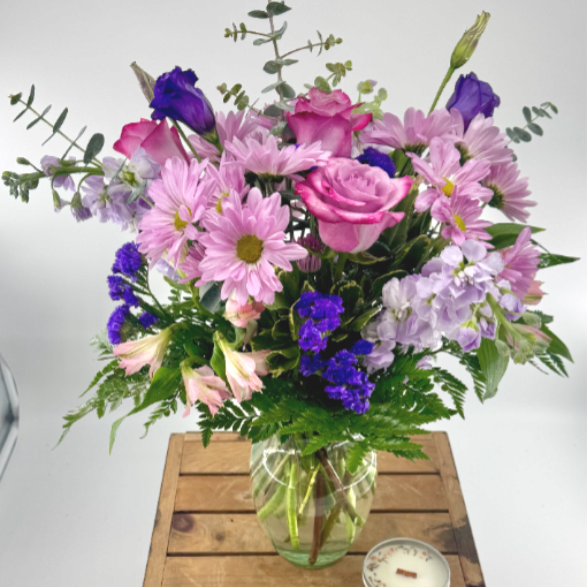 Mixed bouquet of pink daisies, roses, and purple flowers in a glass vase with a small candle beside it.