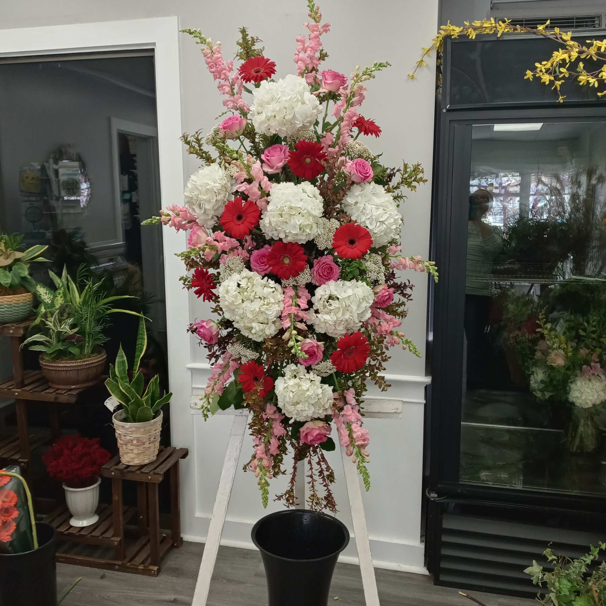 Tall standing spray of white hydrangeas with red gerbera daisies and pink roses on a white easel.