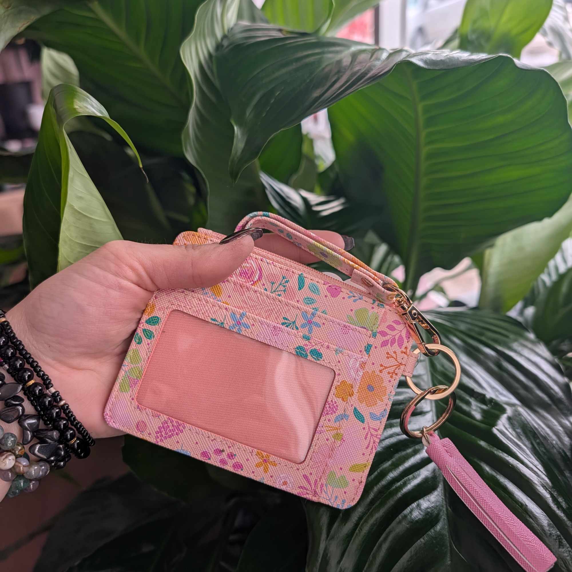 Hand holding a pink floral ID wallet in front of large green leaves.