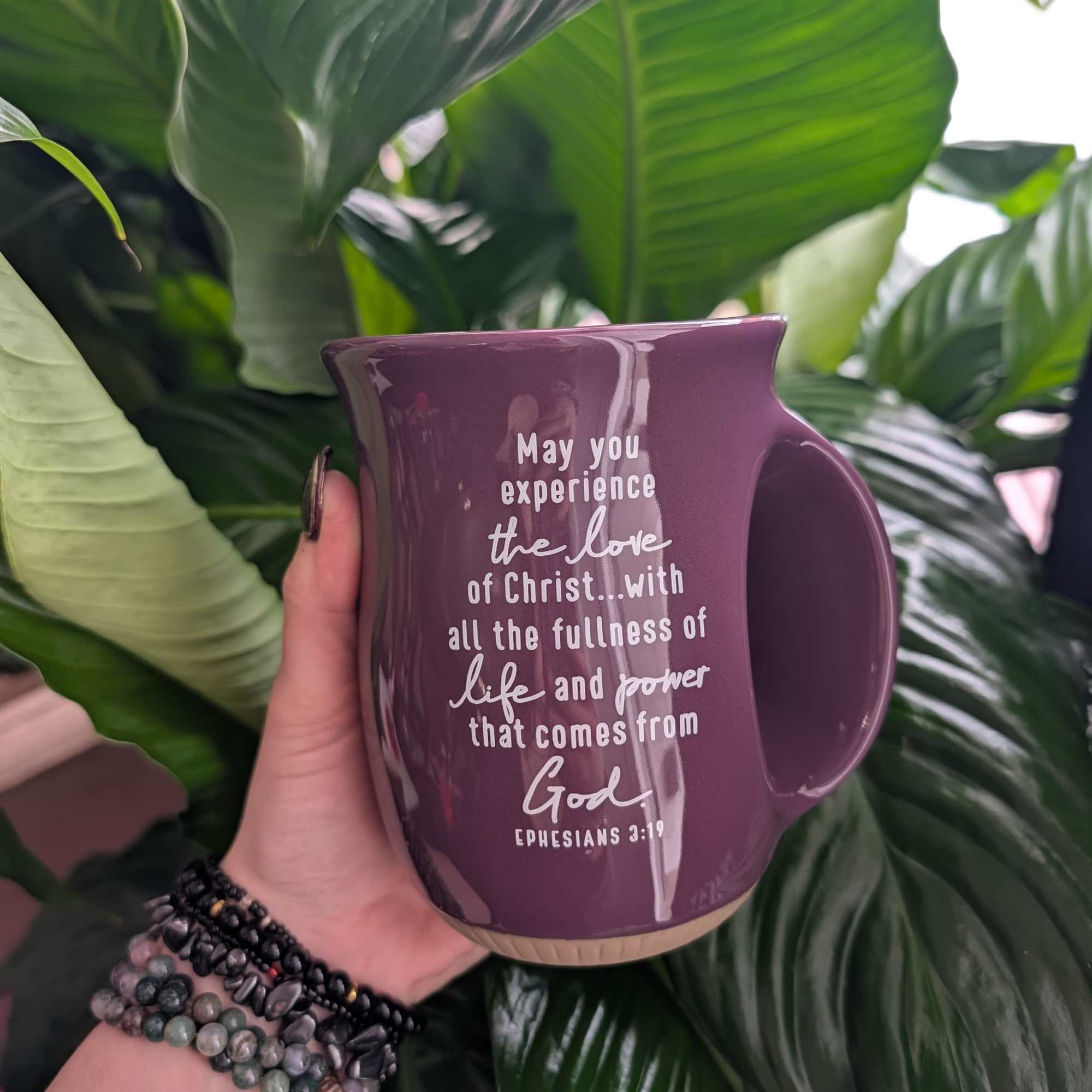 Purple mug with a Bible verse held in front of large green leaves