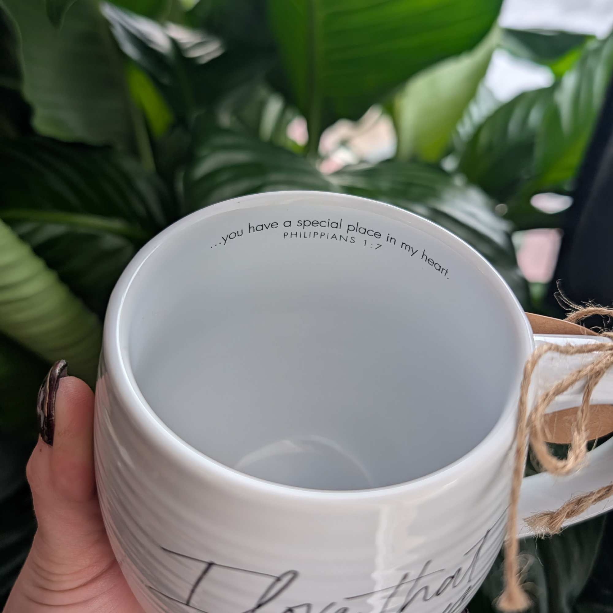 White ceramic mug with a handwritten-style quote, held in front of green leaves.