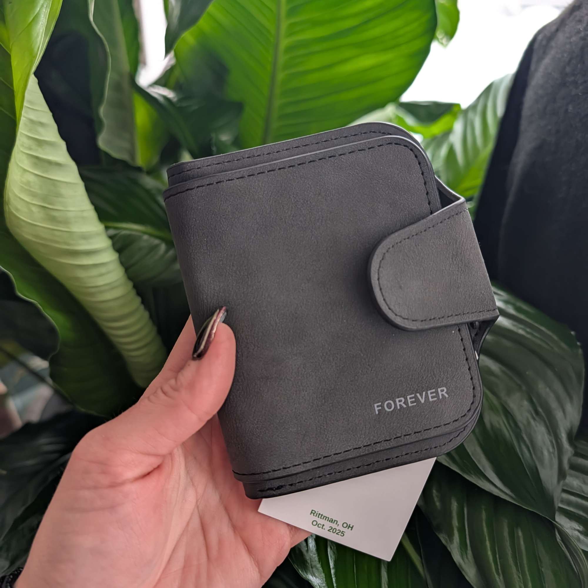 Hand holding a gray wallet in front of large green leaves