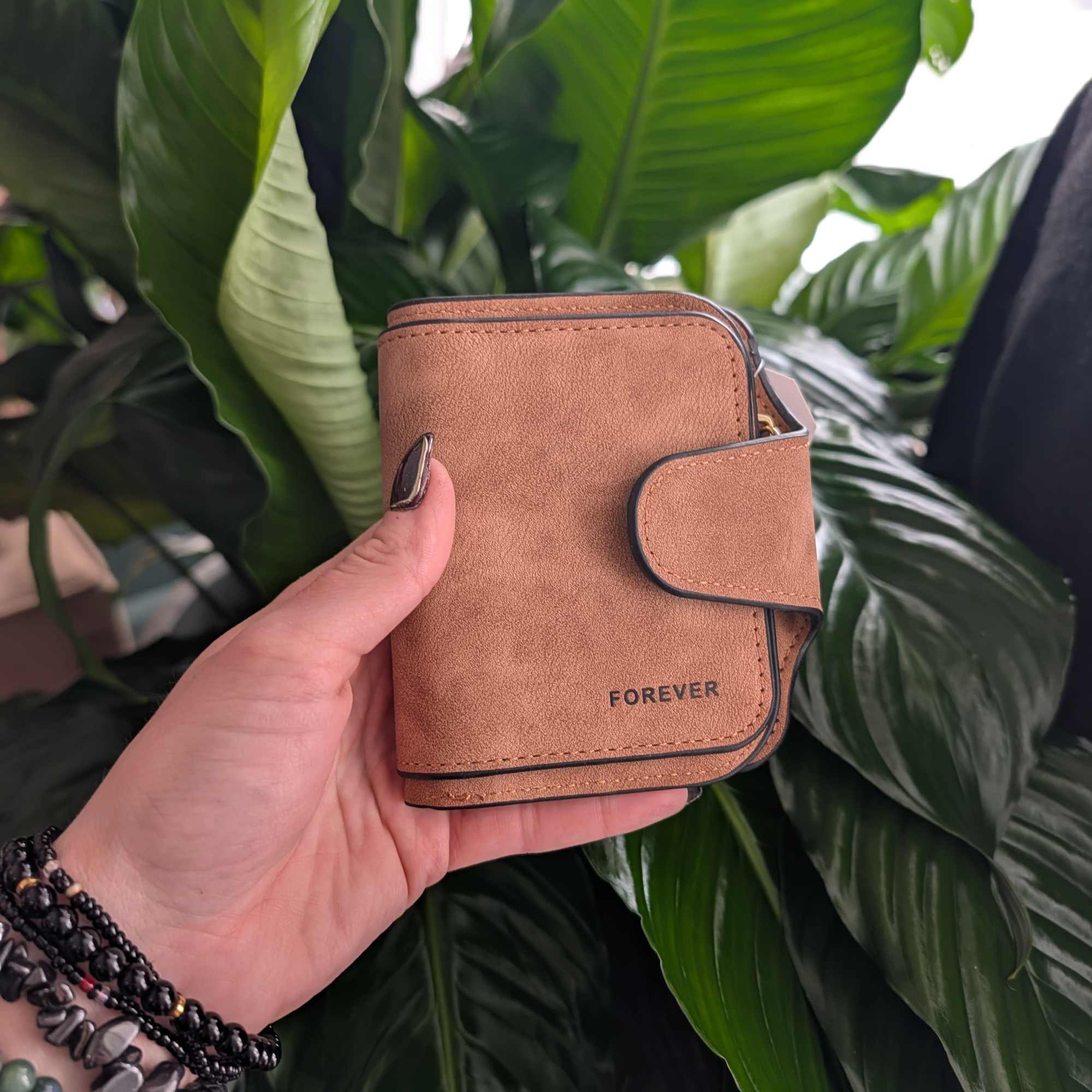 Hand holding a tan wallet in front of large green leaves