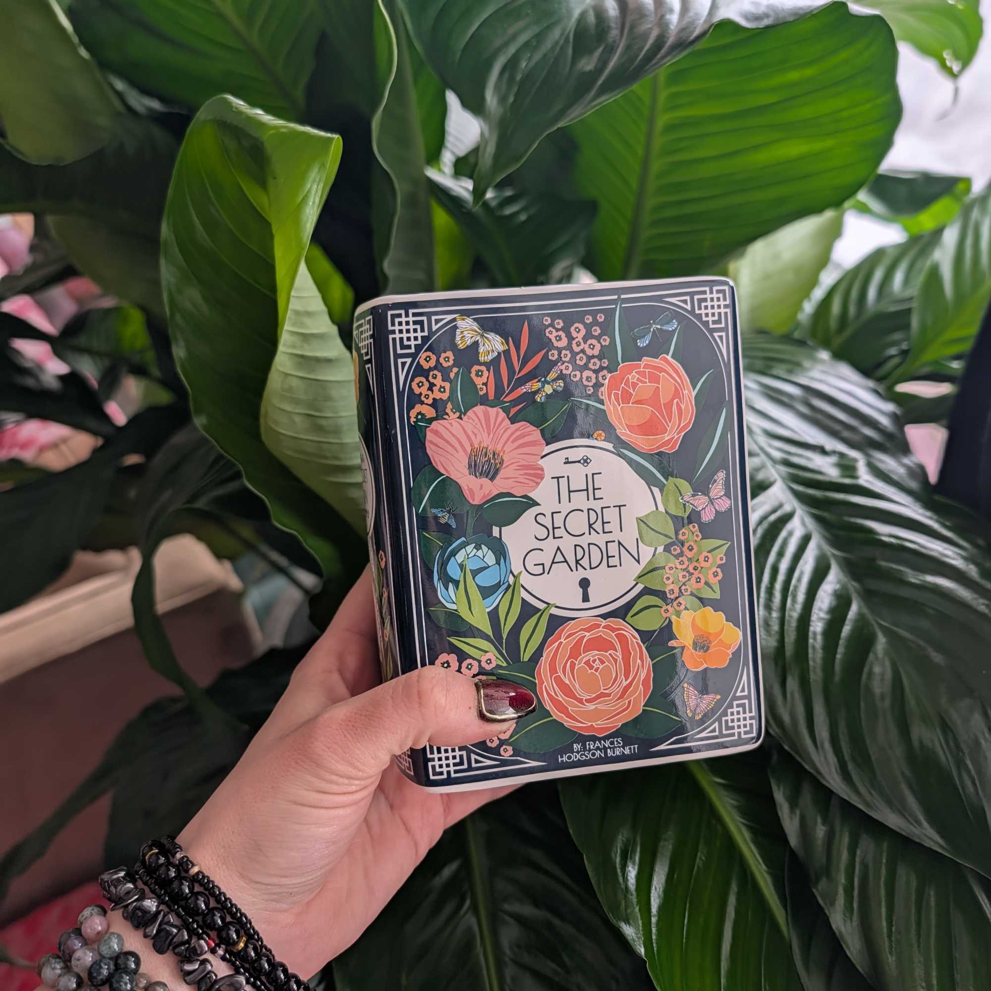 Hand holding a decorative tin box in front of large green leaves