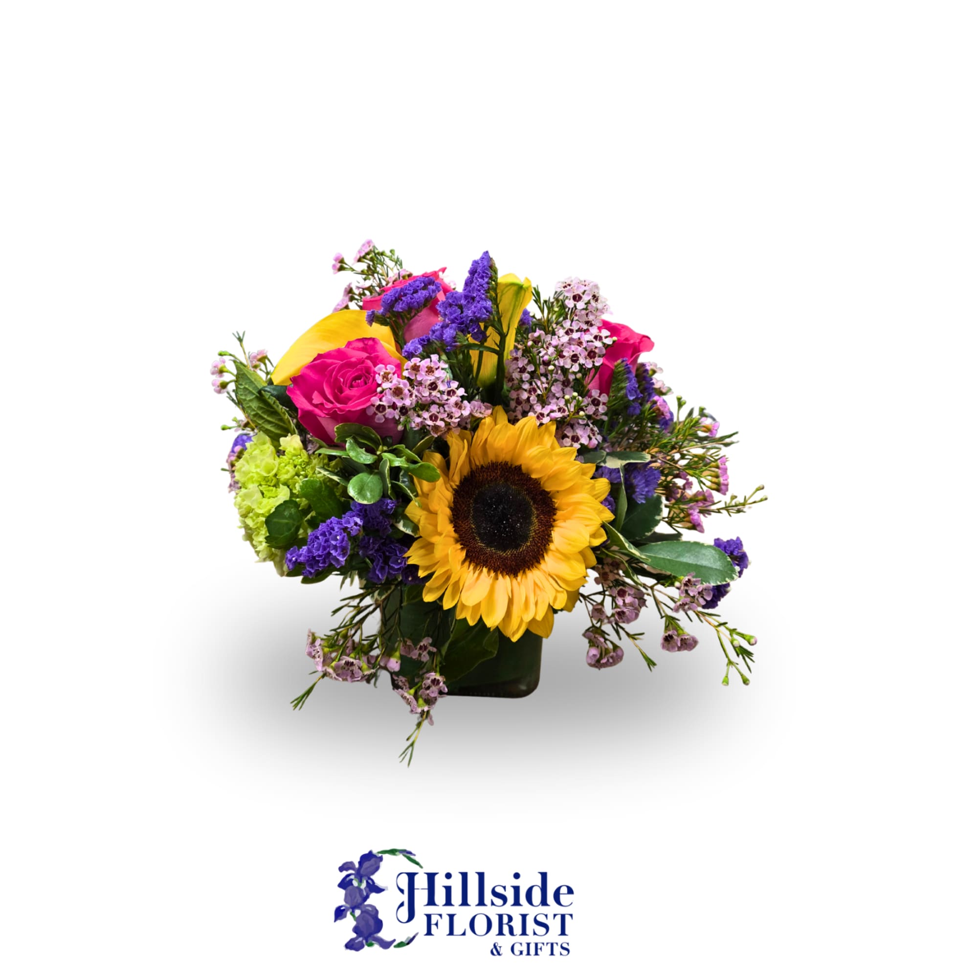 Colorful arrangement with a sunflower, pink roses, yellow calla lilies and purple blooms in a square glass vase