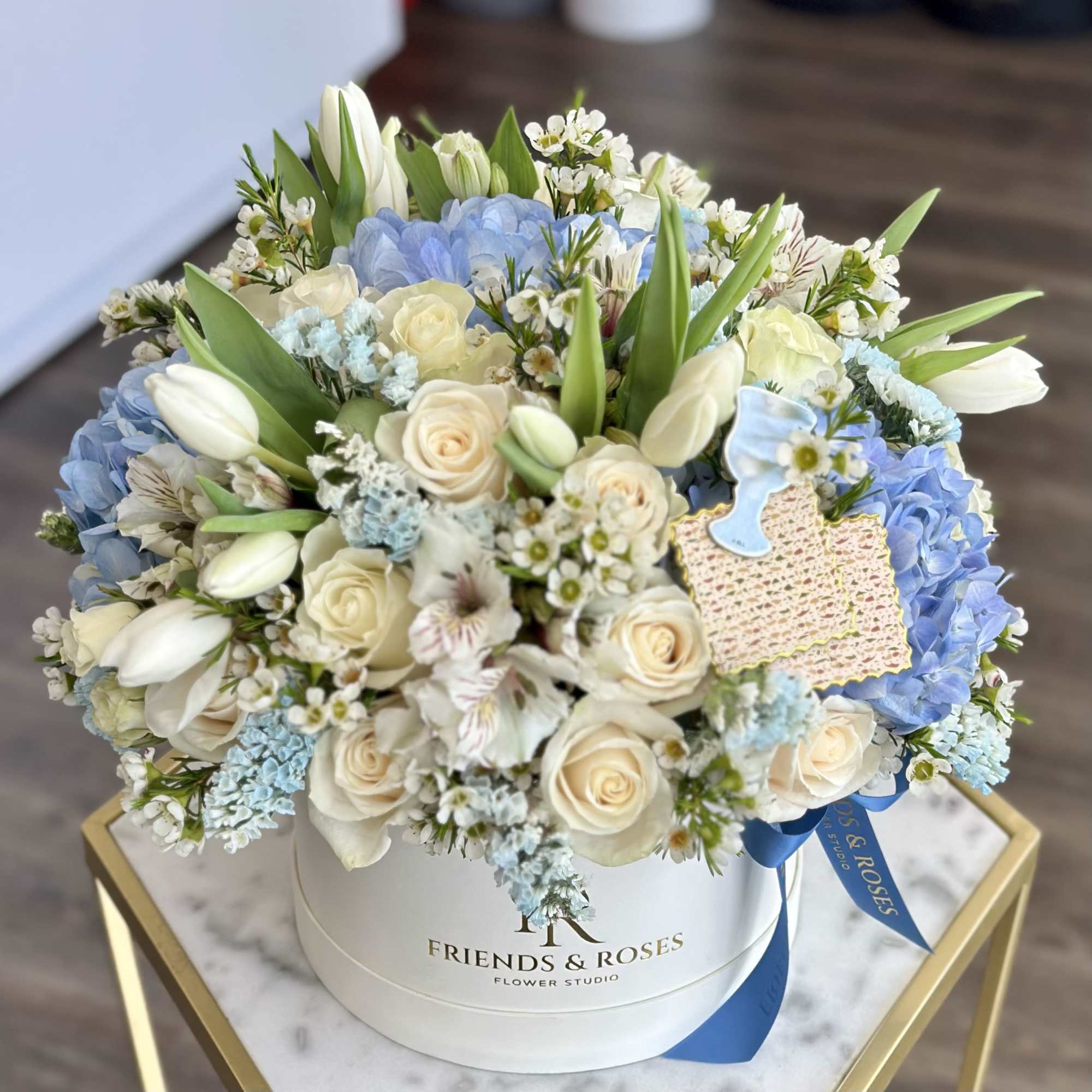 Round hatbox of ivory roses, white tulips, and blue hydrangeas with baby-blue cookie accents.