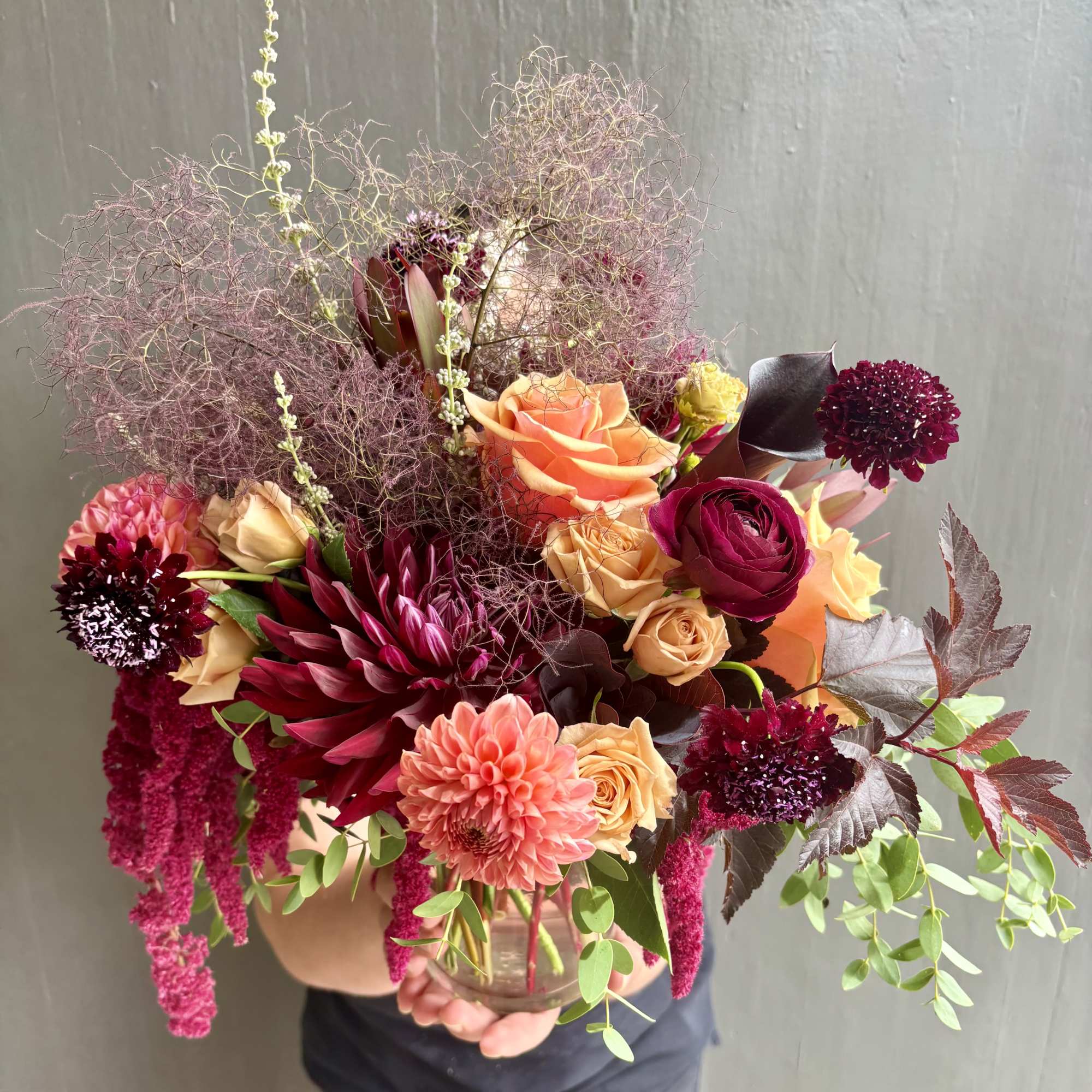 Handheld bouquet of peach, burgundy, and pink flowers in a glass vase