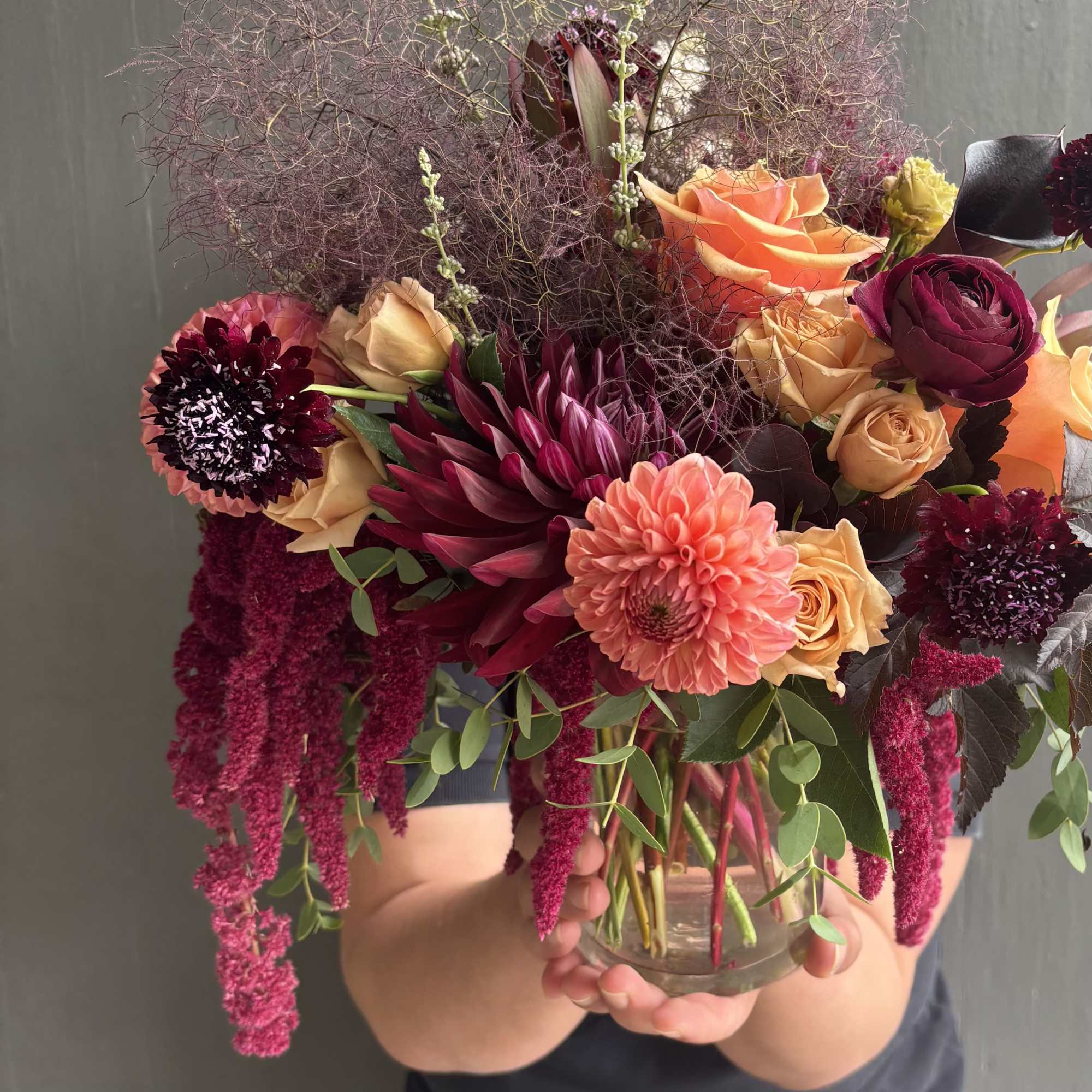 Handheld bouquet of roses, dahlias, and dark burgundy blooms in a glass vase