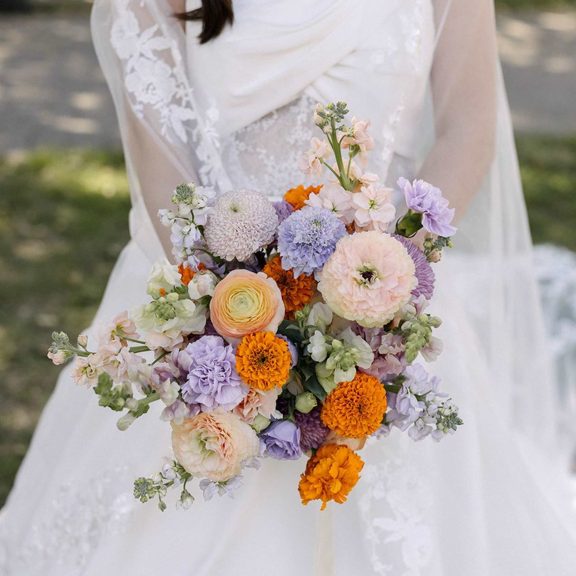 Bridal hand-tied bouquet of peach, orange, and lavender flowers held against a white wedding dress