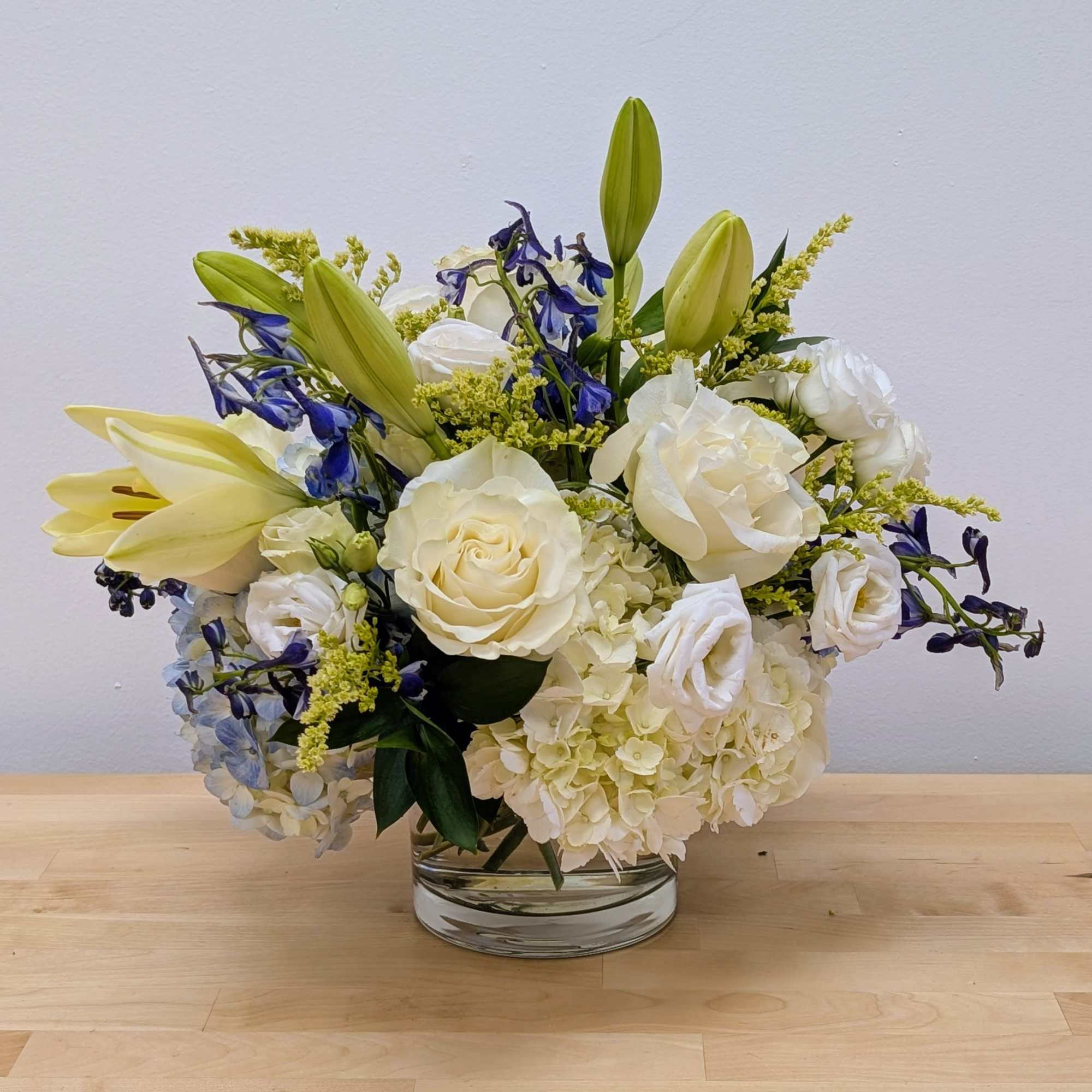 Low glass vase of white roses, hydrangeas, and lilies with blue delphinium accents