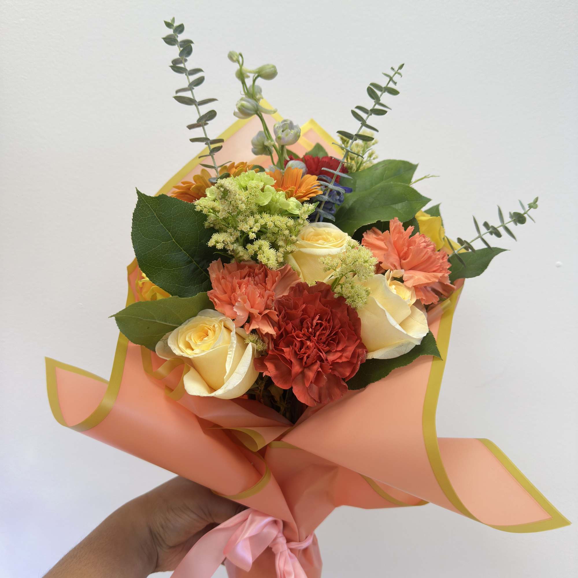 Handheld bouquet of roses, carnations, and orange flowers wrapped in peach paper