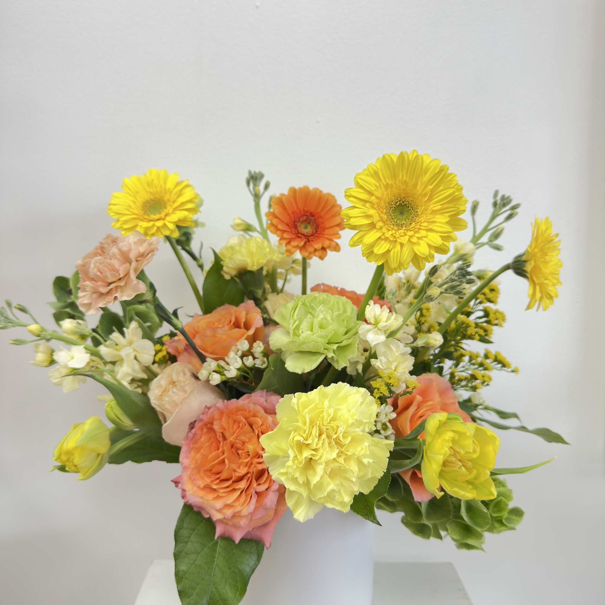 Bright arrangement of yellow gerbera daisies, peach roses, and mixed blooms in a white cylinder vase