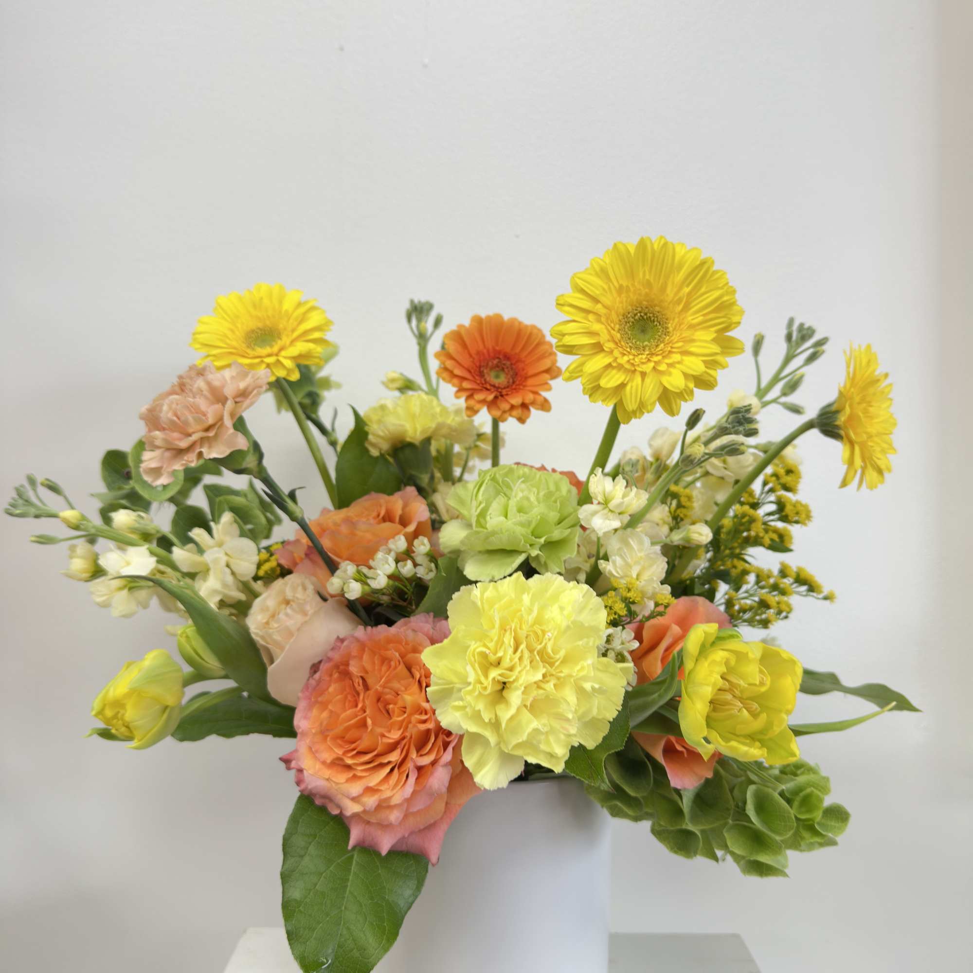 Bright arrangement of yellow and orange gerbera daisies, carnations, and roses in a white vase