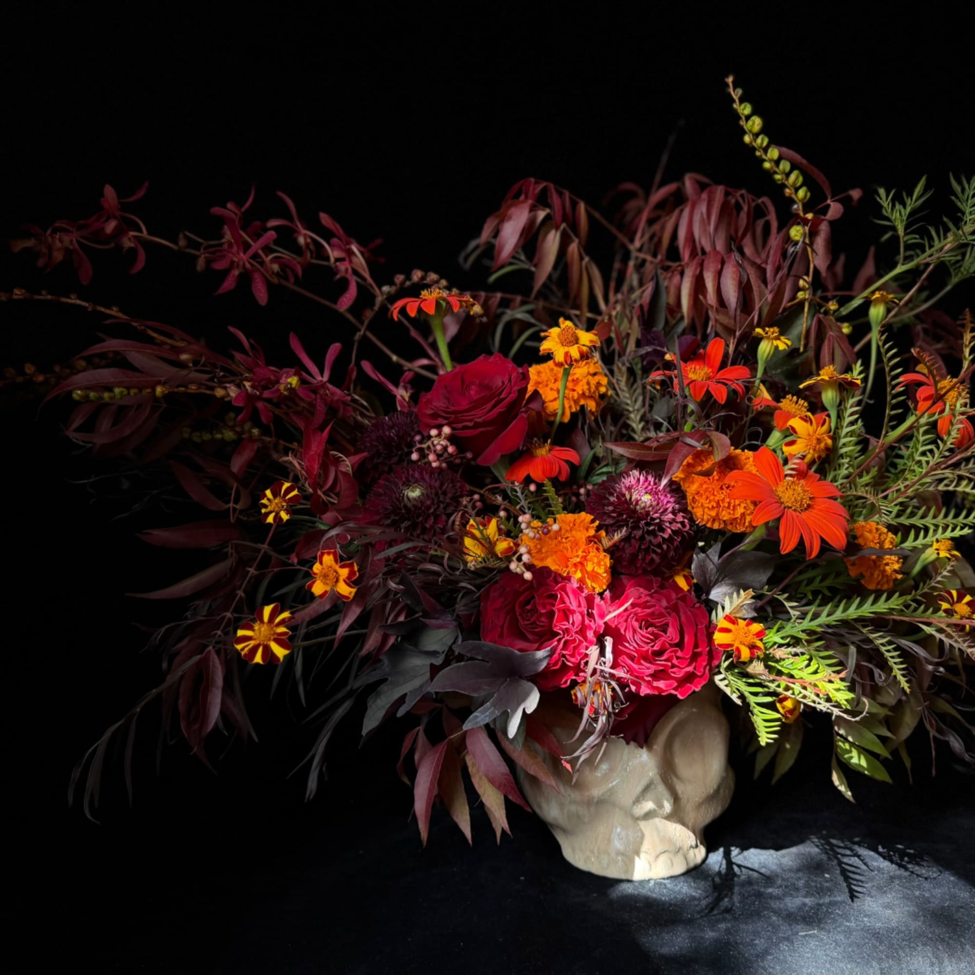 Dark floral arrangement with red roses and orange blooms in a skull-shaped vase
