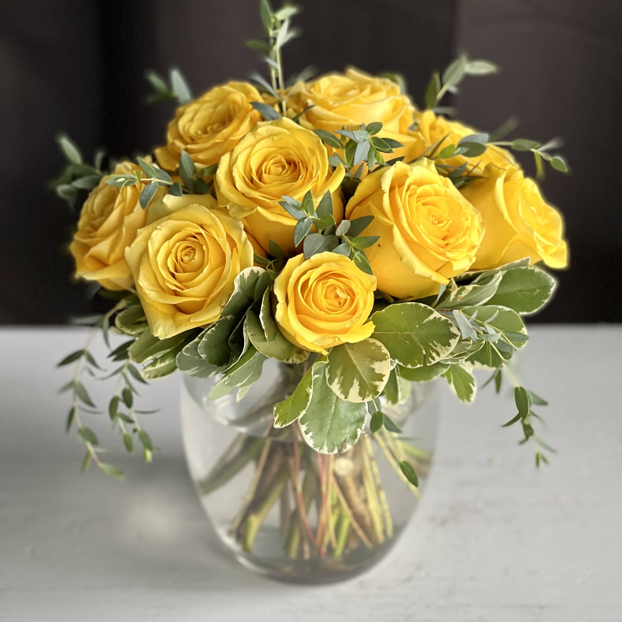 Compact arrangement of yellow roses with variegated foliage in a clear glass vase.