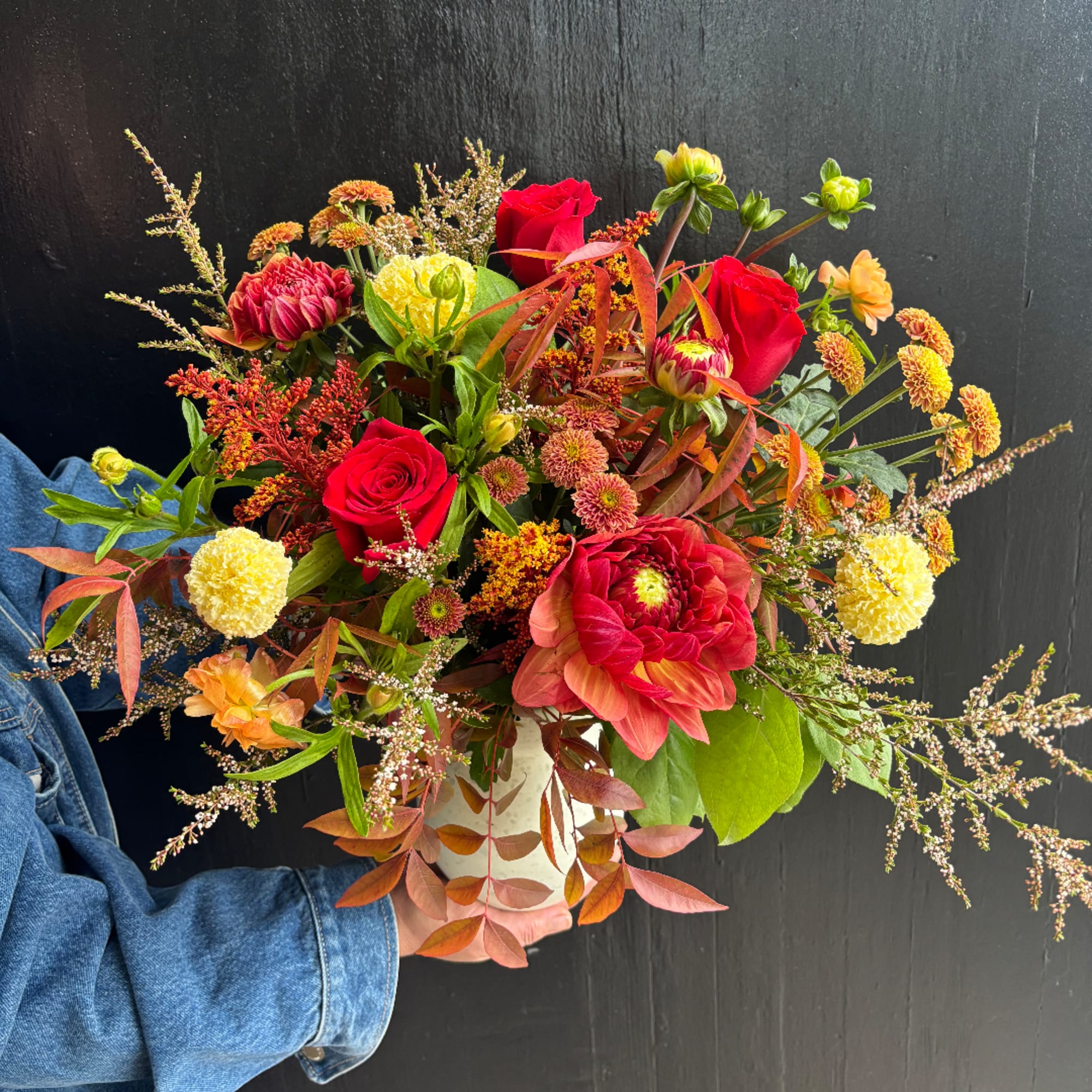 Handheld bouquet of red roses, dahlias, and yellow mums in a white vase