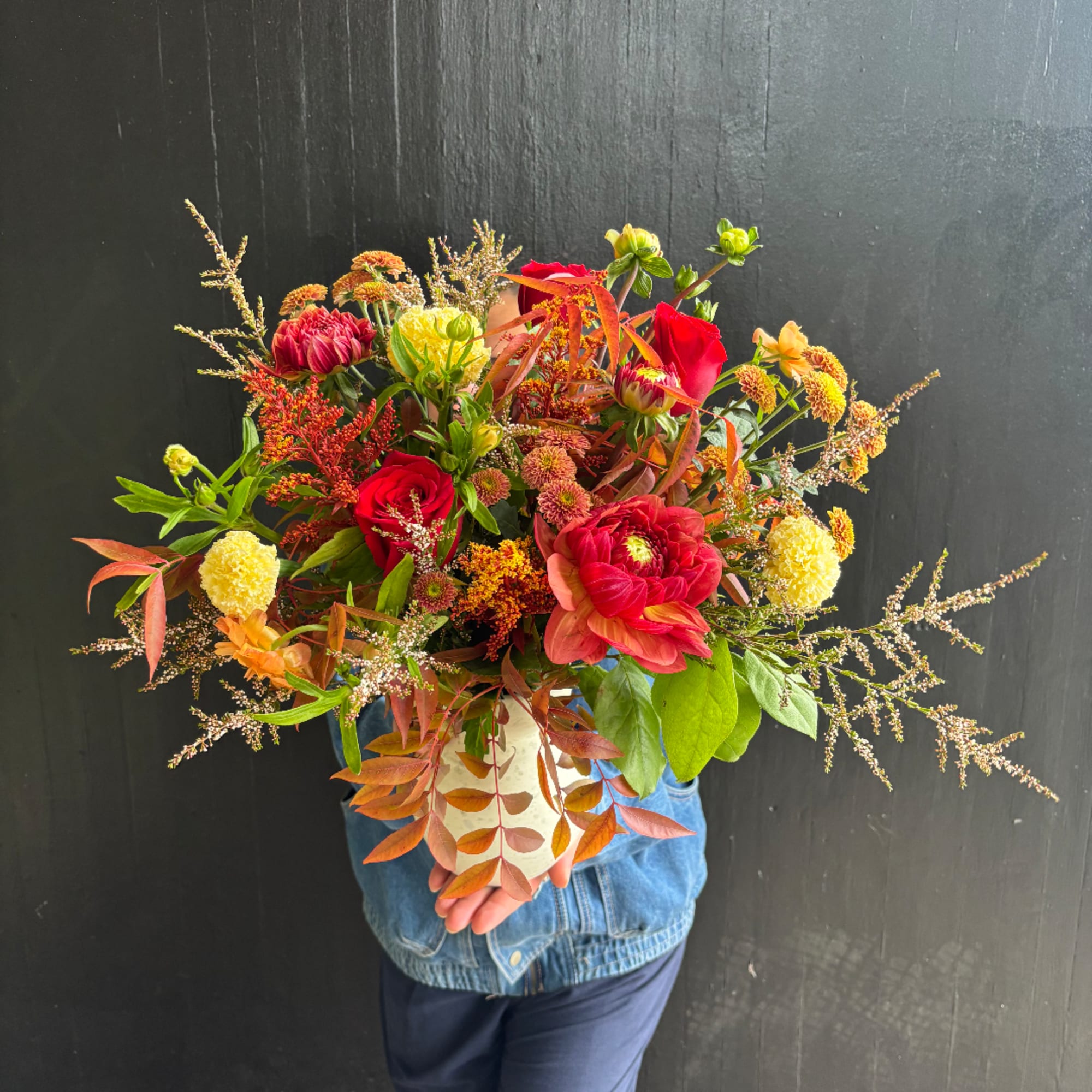 Large mixed bouquet of red, yellow, and orange flowers held in front of a dark wall