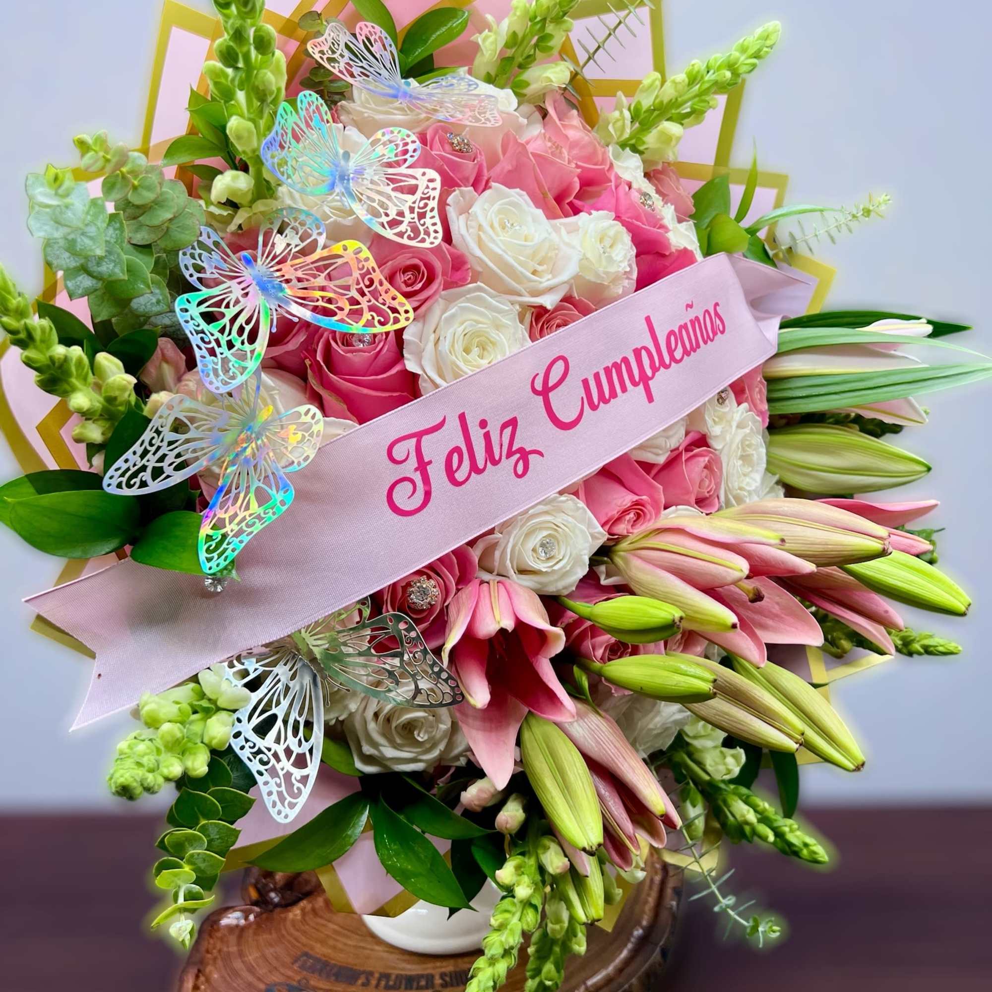 Tall arrangement of pink lilies and pink and white roses with a Feliz Cumpleaños ribbon and butterfly accents.