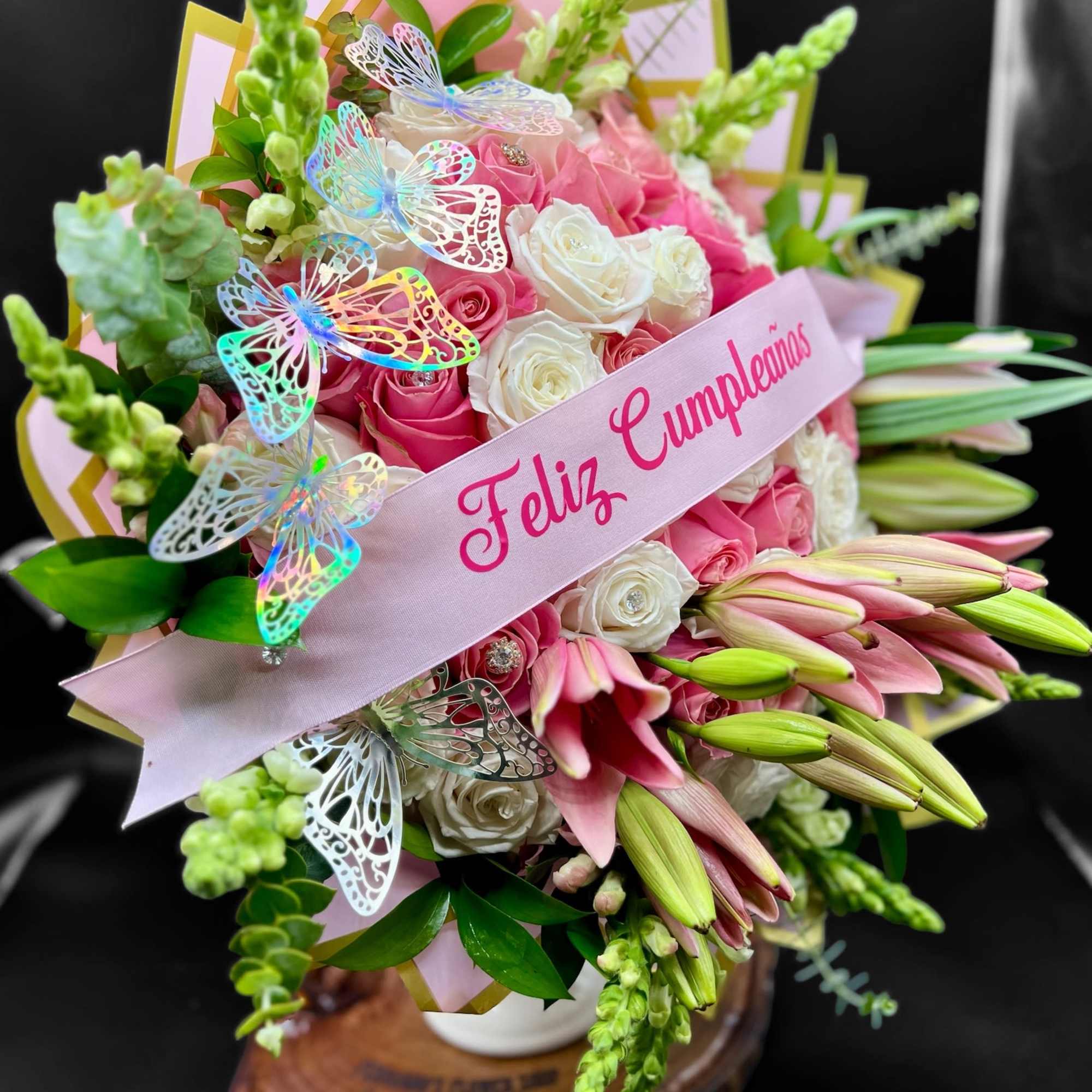 Pink and white birthday flower arrangement with roses, lilies, and butterflies around a Feliz Cumpleaños ribbon.